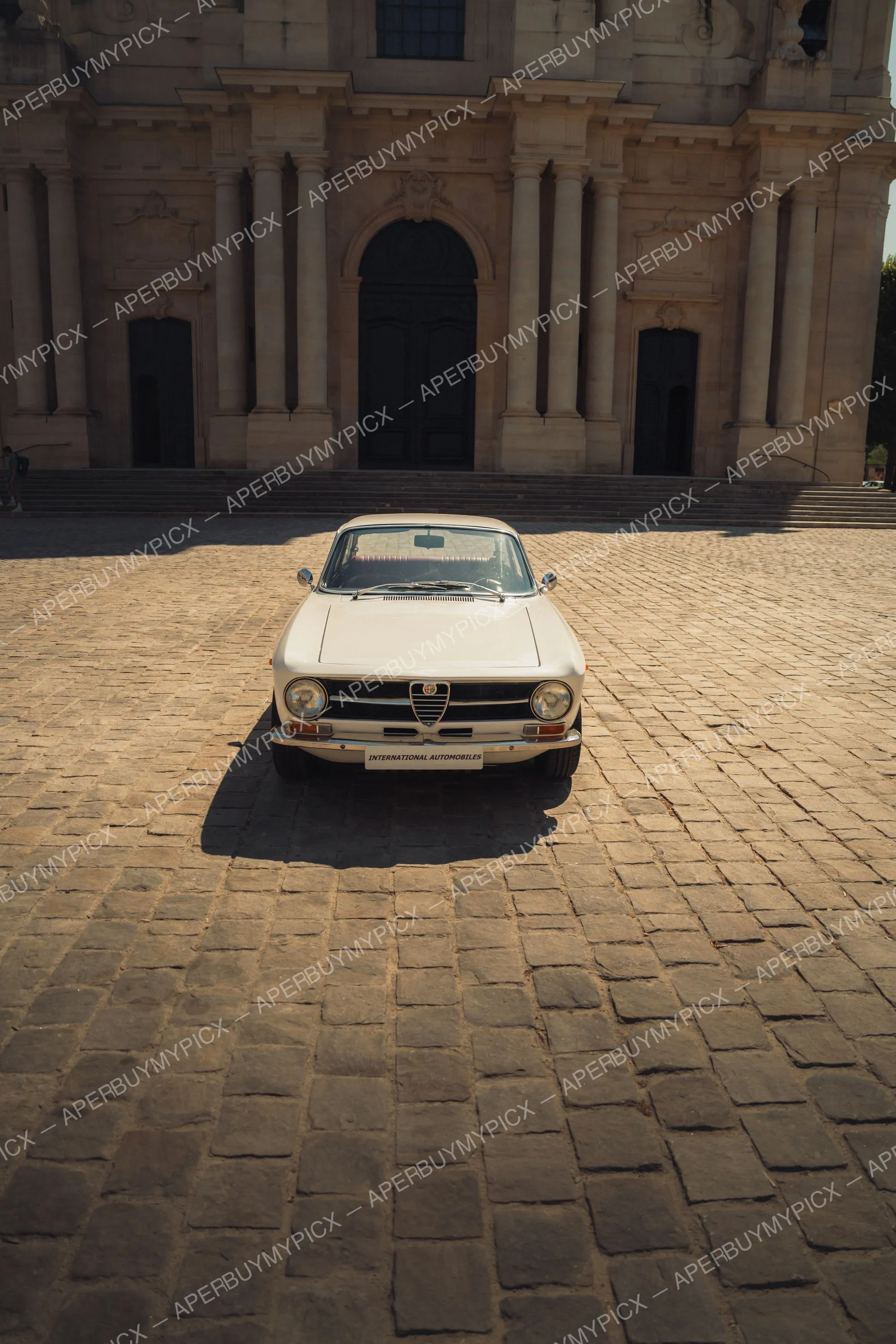 Alfa Romeo GT 1600 crème vue de face, garée sur une place pavée devant une façade baroque en pierre avec de hautes colonnes et une porte cintrée sombre, sous une lumière du jour éclatante.