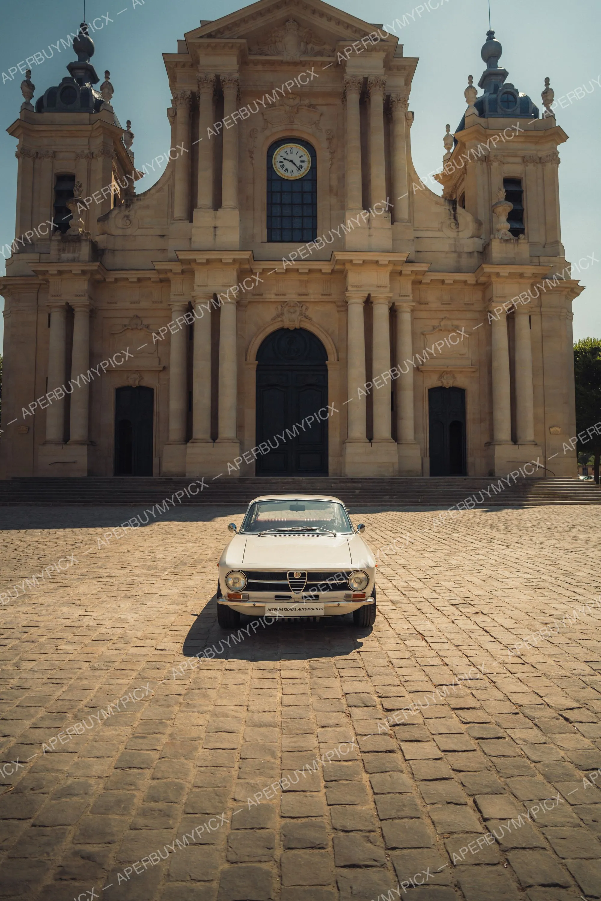 Alfa Romeo GT 1600 de couleur crème vue de face, stationnée sur une place pavée devant une majestueuse cathédrale baroque avec horloge et deux tours.