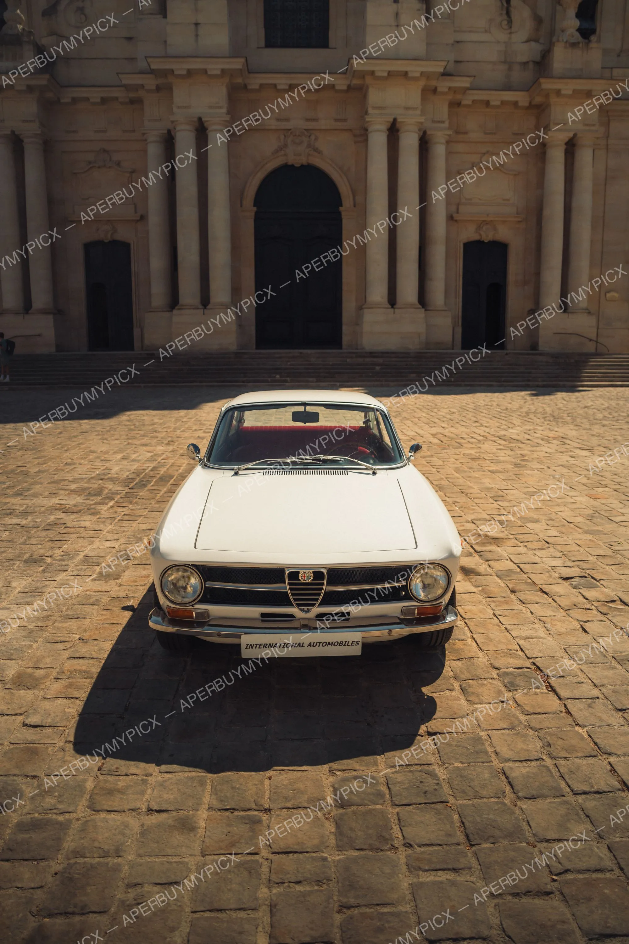 Alfa Romeo GT 1600 blanche vue de face, garée sur une place pavée devant une imposante façade baroque en pierre avec portail voûté.