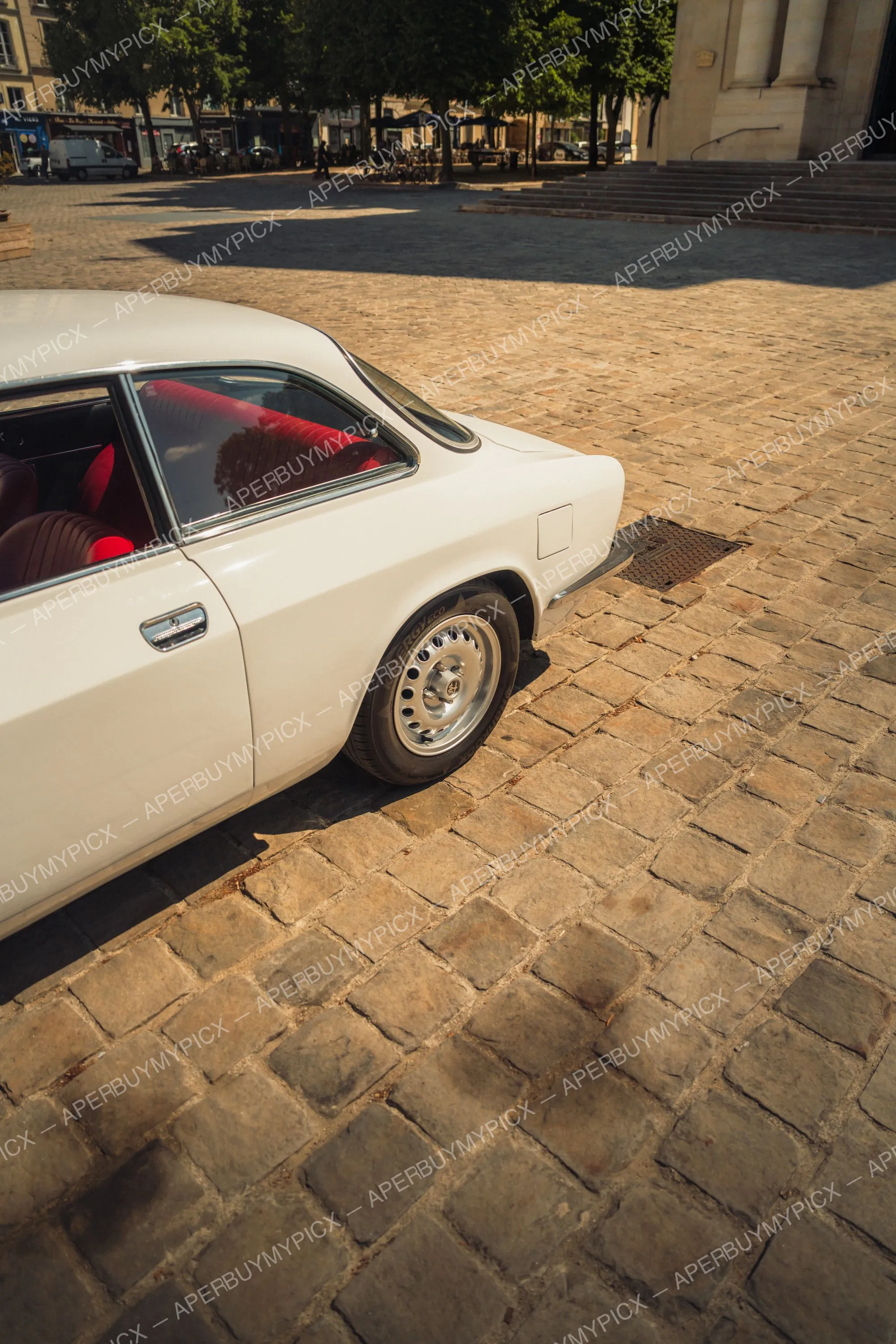 Vue arrière d'une Alfa Romeo 1600 blanche vintage à l'intérieur rouge, garée sur une place pavée ensoleillée avec arbres et bâtiments en pierre en arrière-plan.