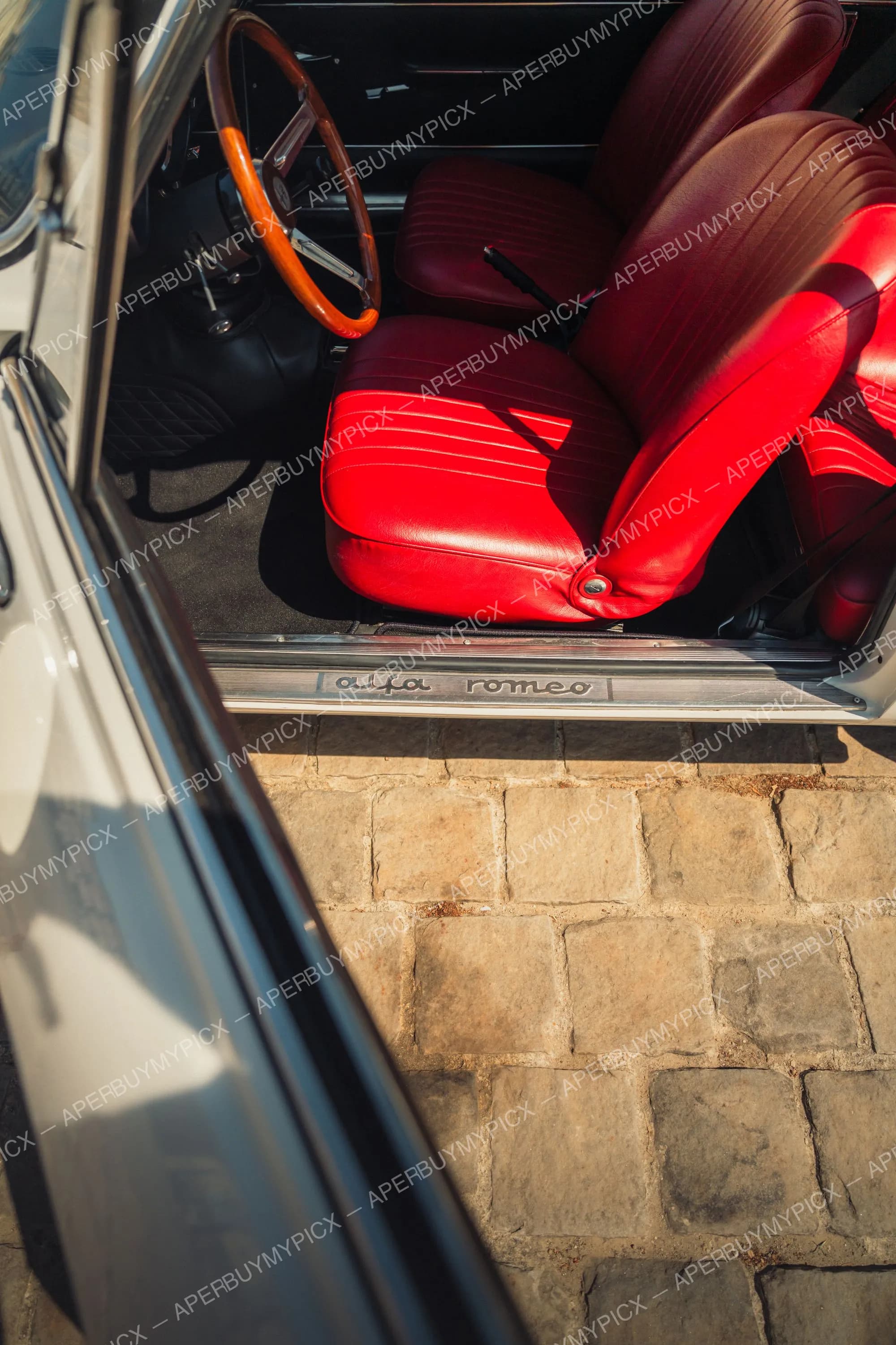 Intérieur d'une Alfa Romeo 1600 vintage avec sièges en cuir rouge, volant en bois, seuil de porte gravé 'alfa romeo', vue par la portière ouverte sur des pavés ensoleillés.