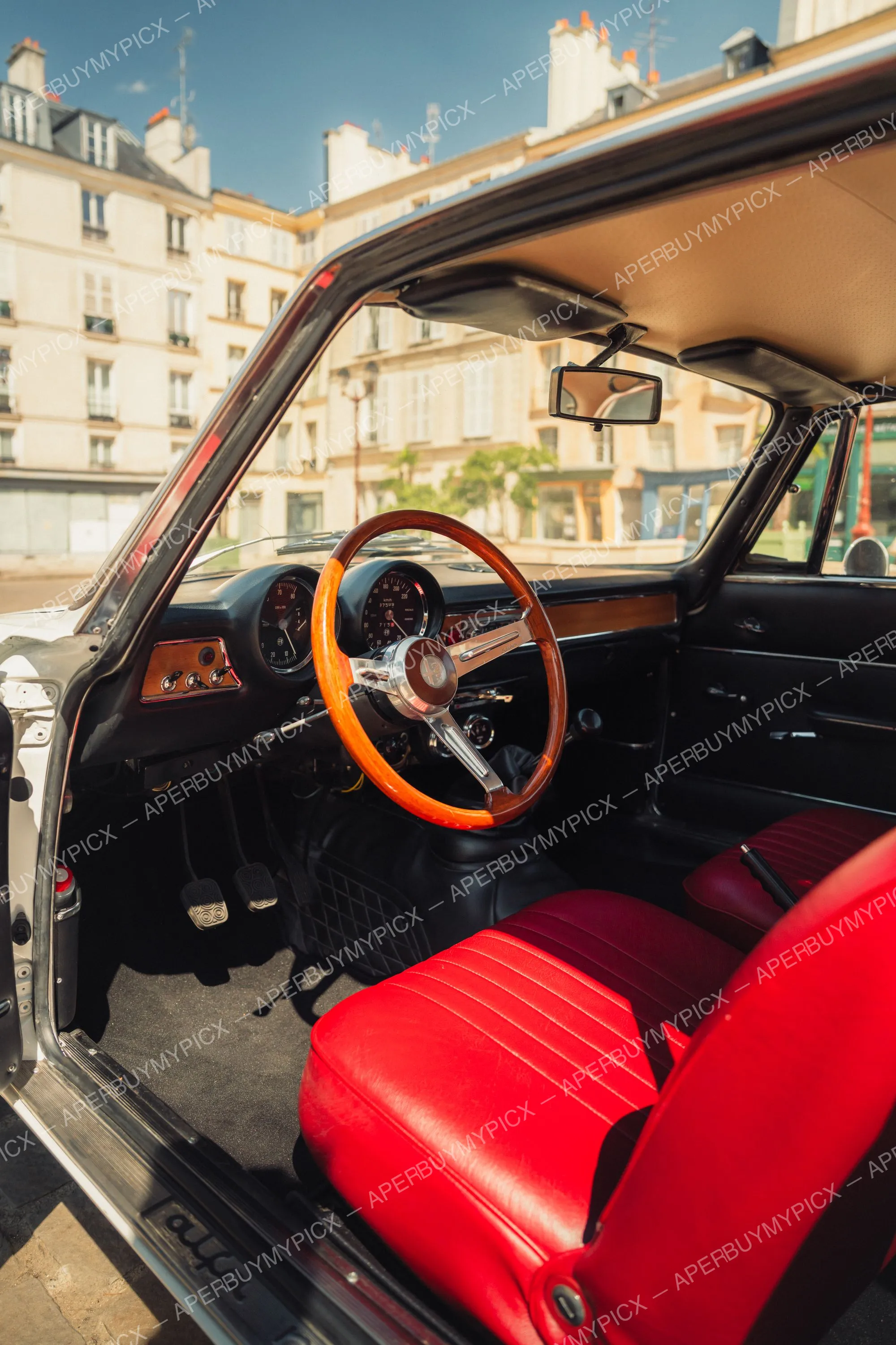 Intérieur d'une Alfa Romeo 1600 classique avec volant en bois, sièges en cuir rouge et tableau de bord vintage, garée dans une rue parisienne ensoleillée.