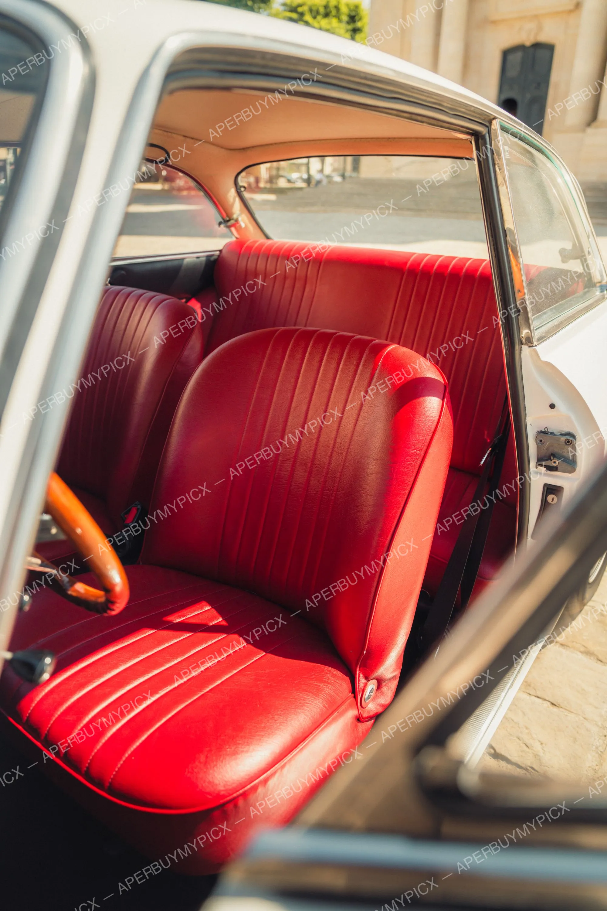Intérieur d'une Alfa Romeo 1600 vintage avec sièges en cuir rouge nervuré, volant en bois et carrosserie blanche, vu à travers la portière ouverte au soleil.
