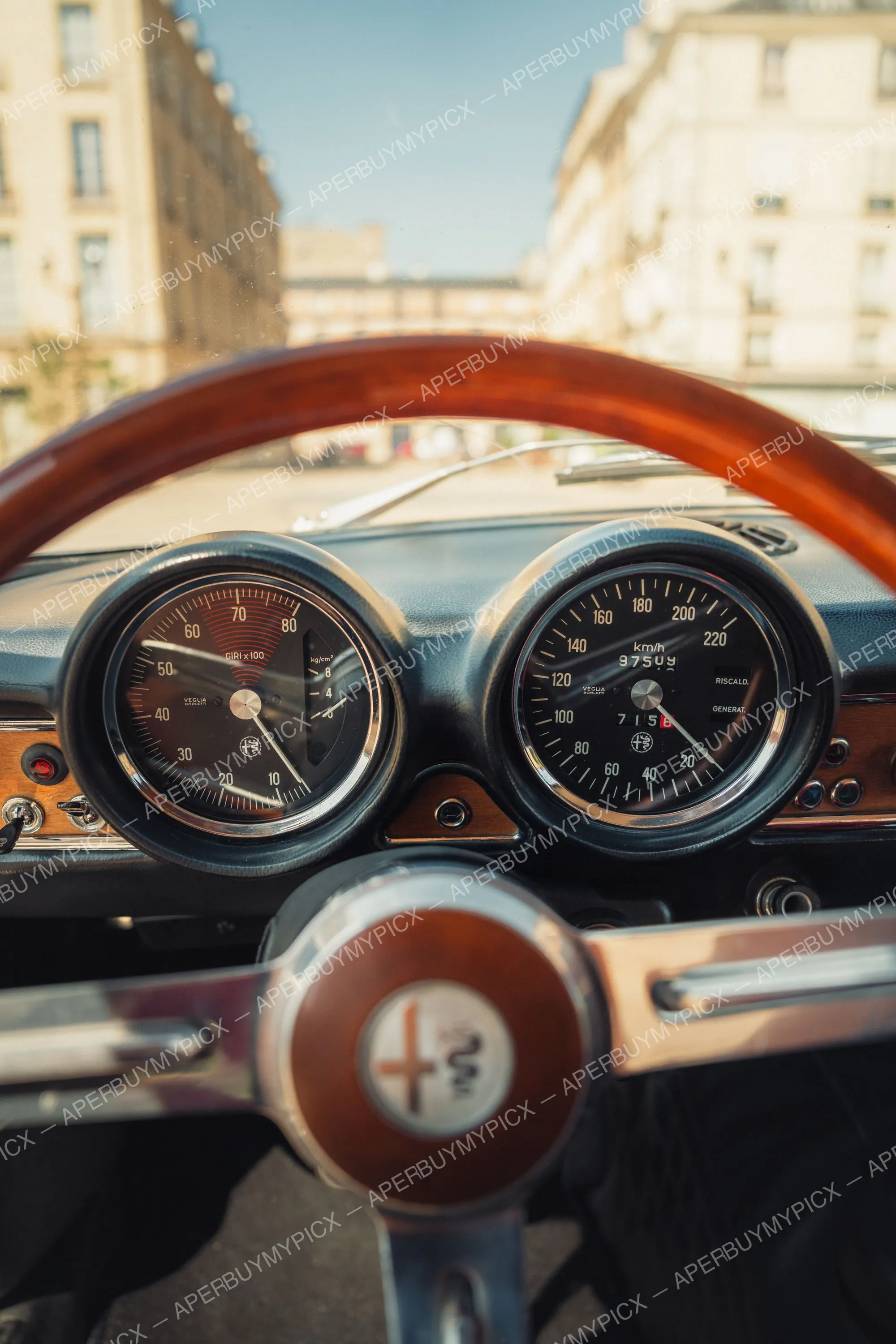 Vue du tableau de bord d'une Alfa Romeo 1600 vintage à travers un volant en bois, montrant le compte-tours Veglia Borletti et le compteur de vitesse km/h affichant 97509 km, avec une rue parisienne floue en arrière-plan.