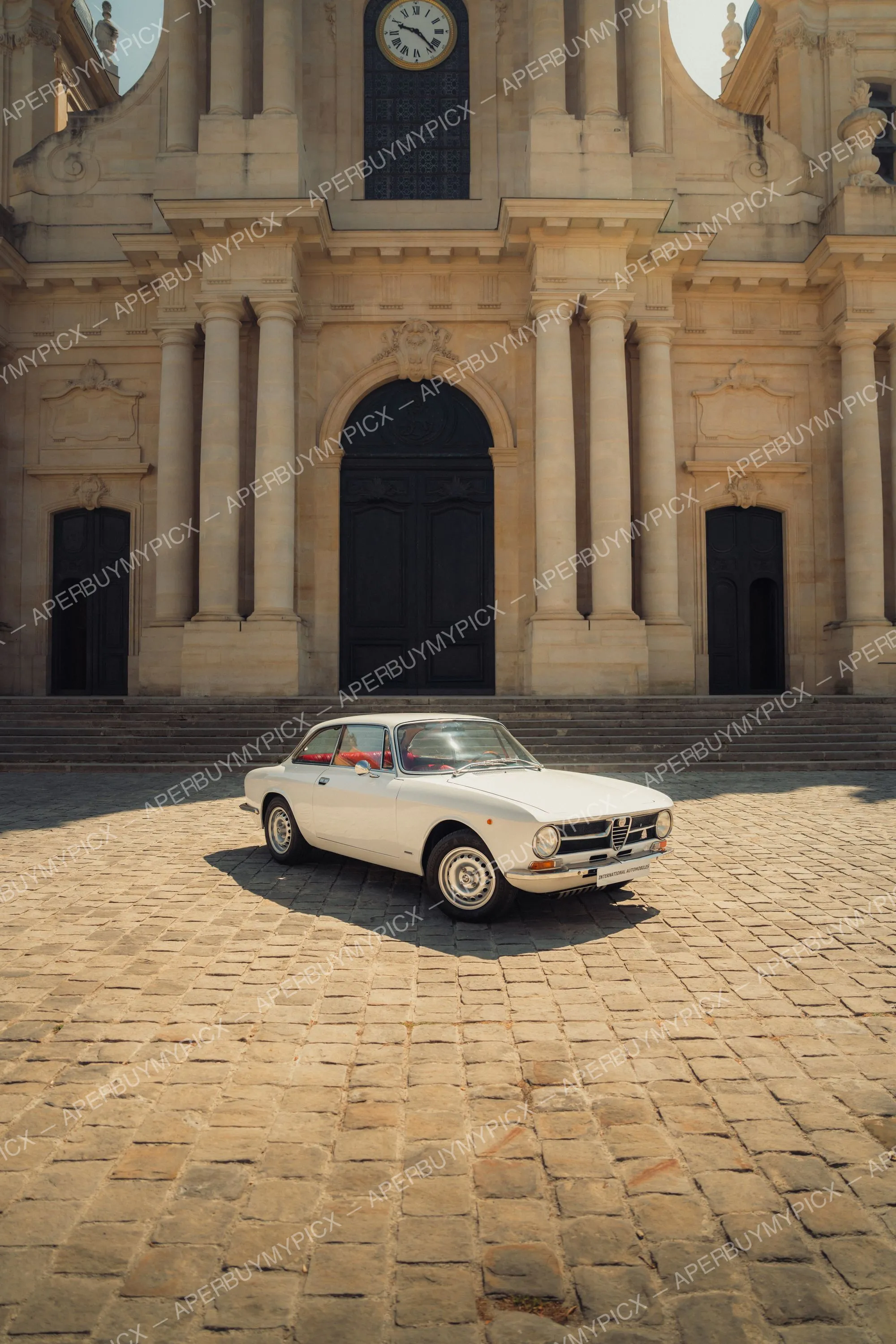 Alfa Romeo 1600 GT blanche vintage garée sur une place pavée devant une imposante église baroque en pierre avec colonnes, grande porte noire et horloge.