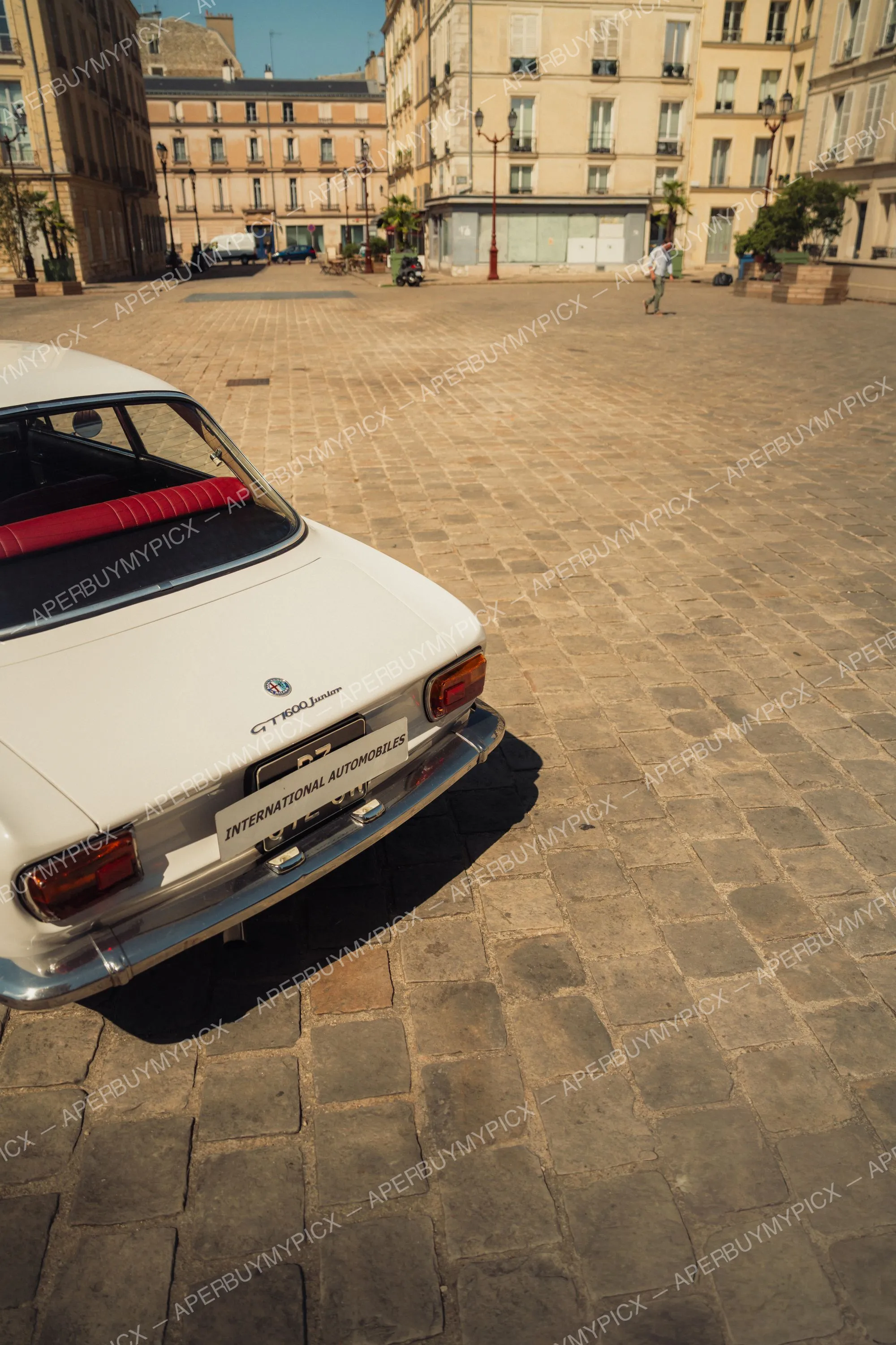 Vue arrière d'une Alfa Romeo GT 1600 Junior blanche à sièges rouges, garée sur une place pavée parisienne bordée d'immeubles haussmanniens.
