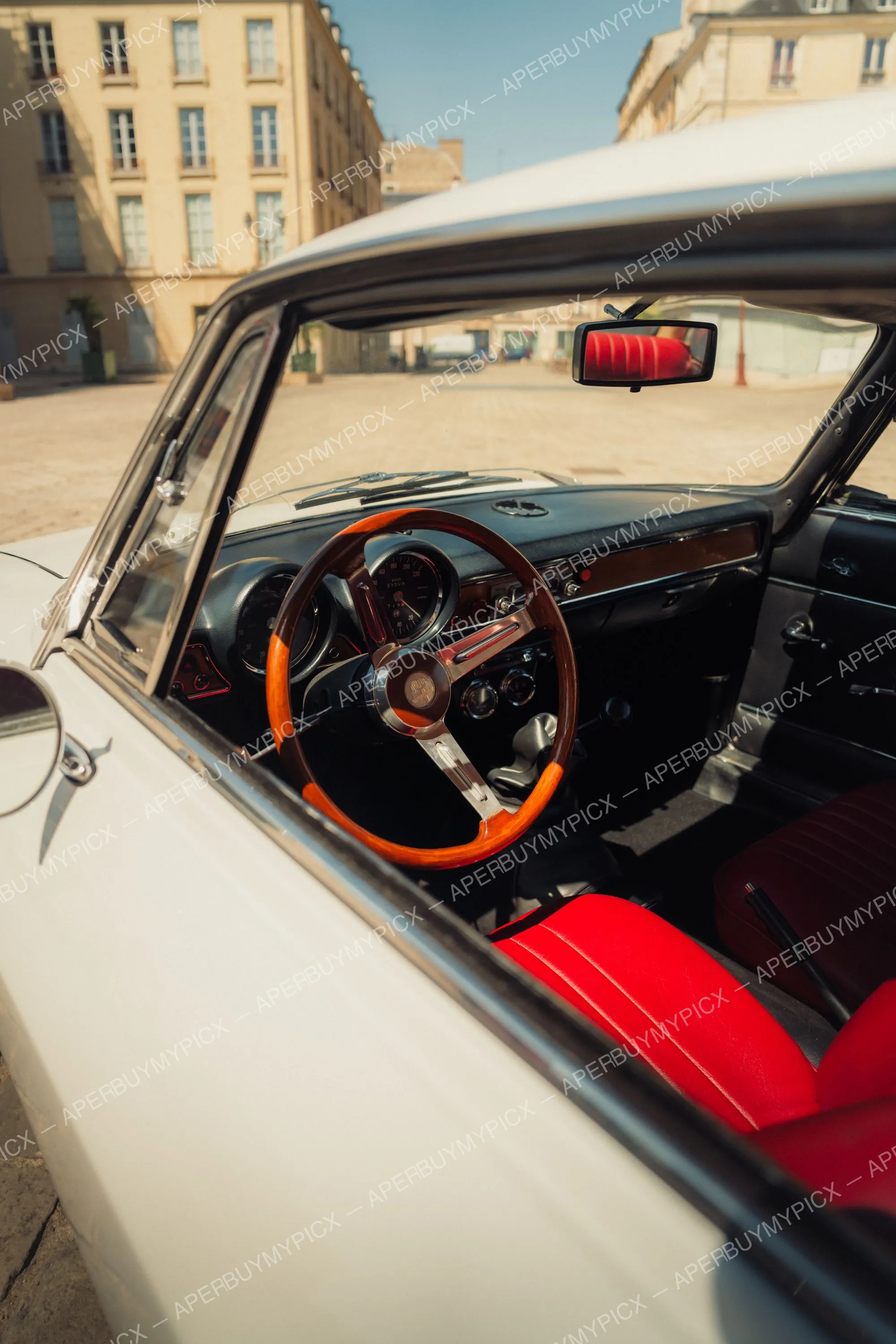 Intérieur d'une Alfa Romeo 1600 blanche avec volant en bois, sièges en cuir rouge et tableau de bord vintage, garée dans une rue ensoleillée.