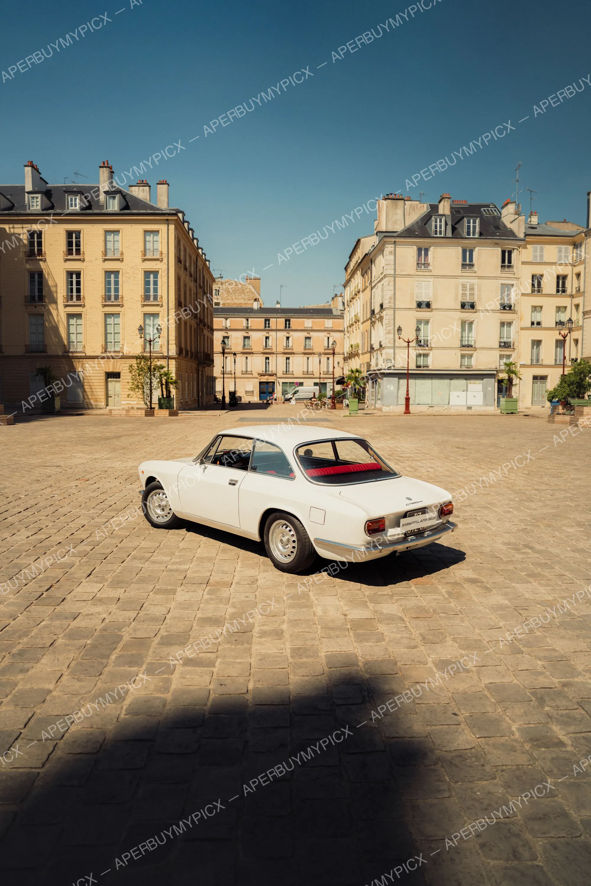 Alfa Romeo GT 1600 blanche vue de trois quarts arrière, garée sur une place pavée entourée d'immeubles haussmanniens sous un ciel bleu.