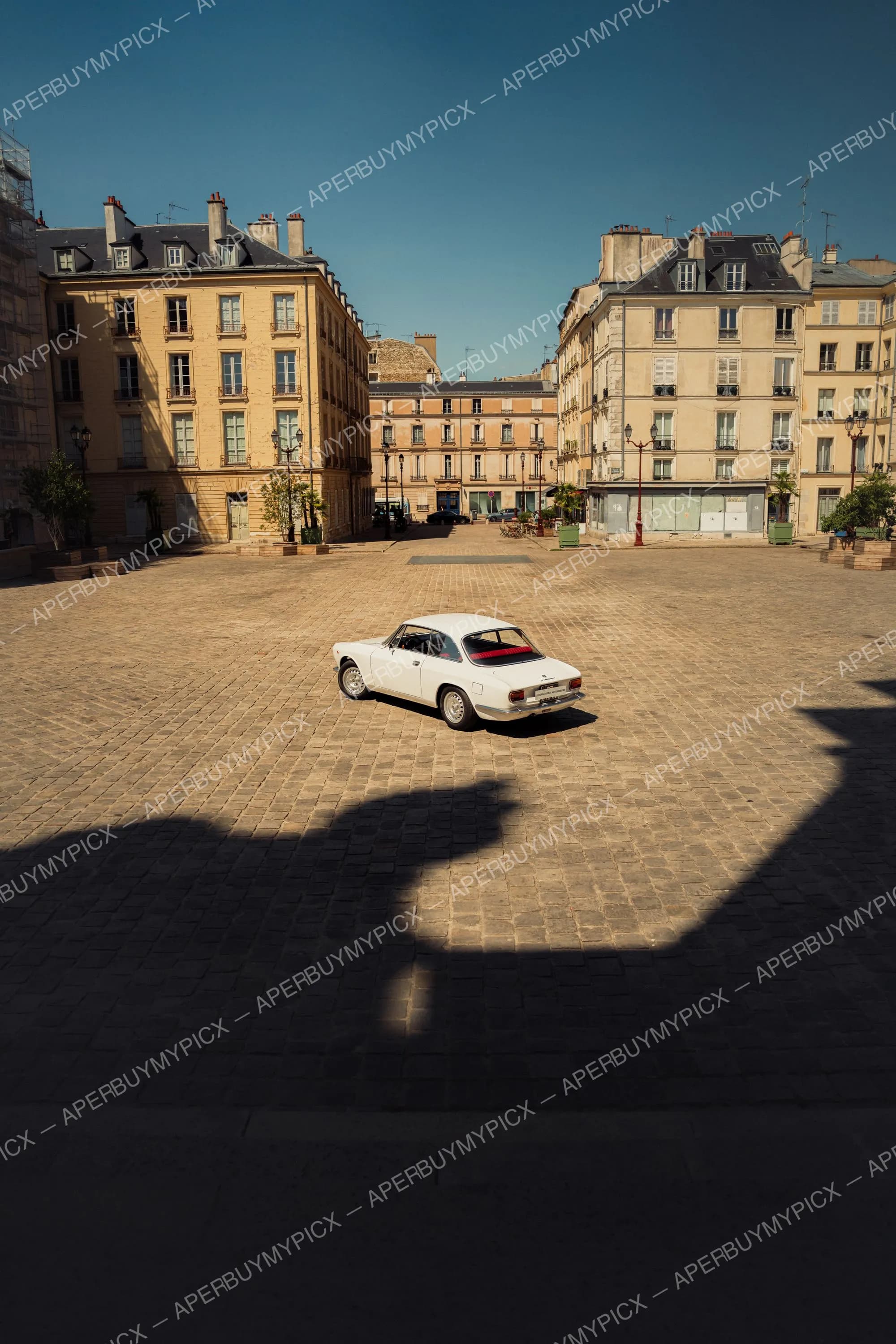 Alfa Romeo GT 1600 blanche stationnée au centre d'une place pavée bordée d'immeubles haussmanniens sous un ciel bleu ensoleillé