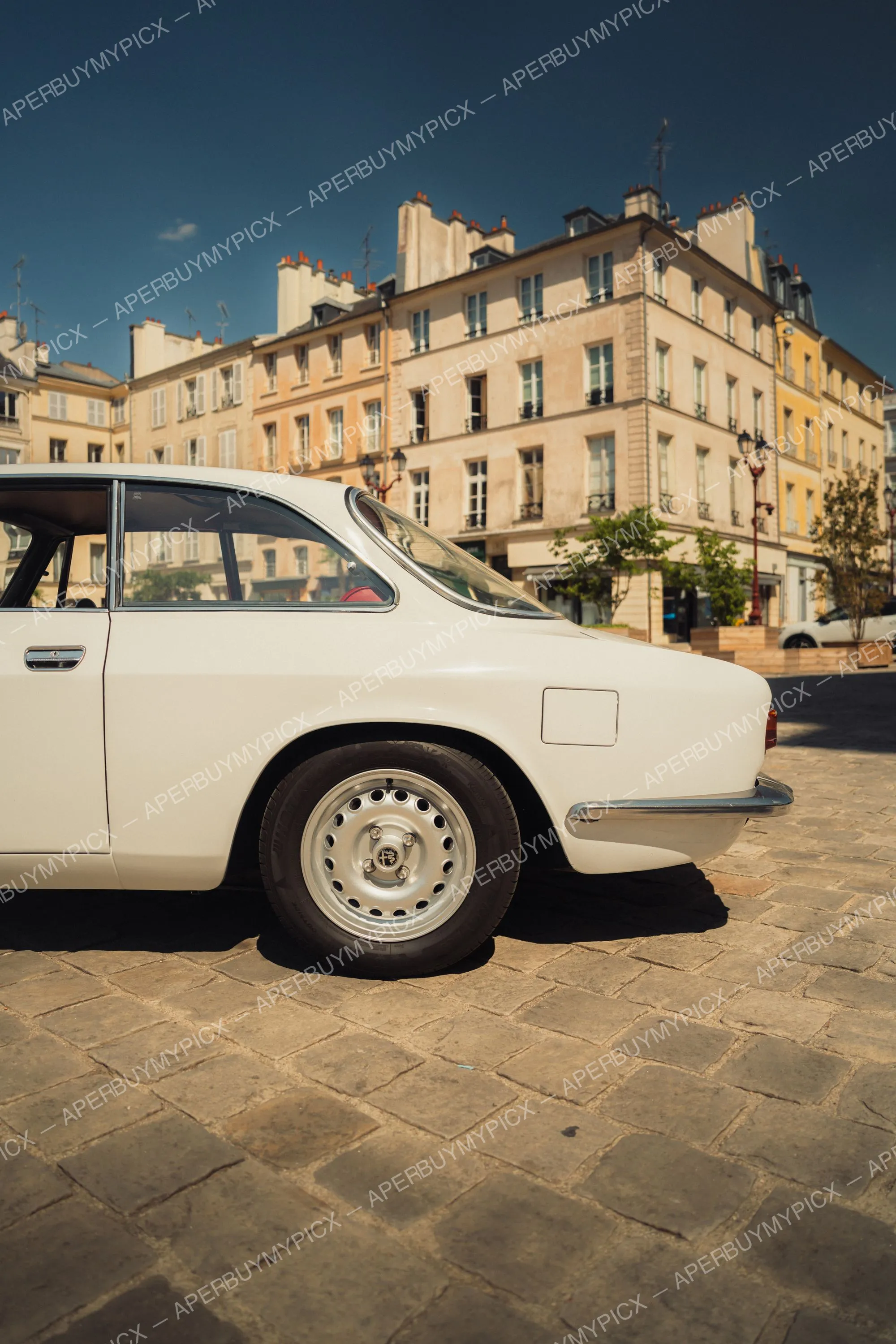 Alfa Romeo 1600 GT blanche vue de profil arrière, garée sur des pavés devant des immeubles haussmanniens parisiens sous un ciel bleu.