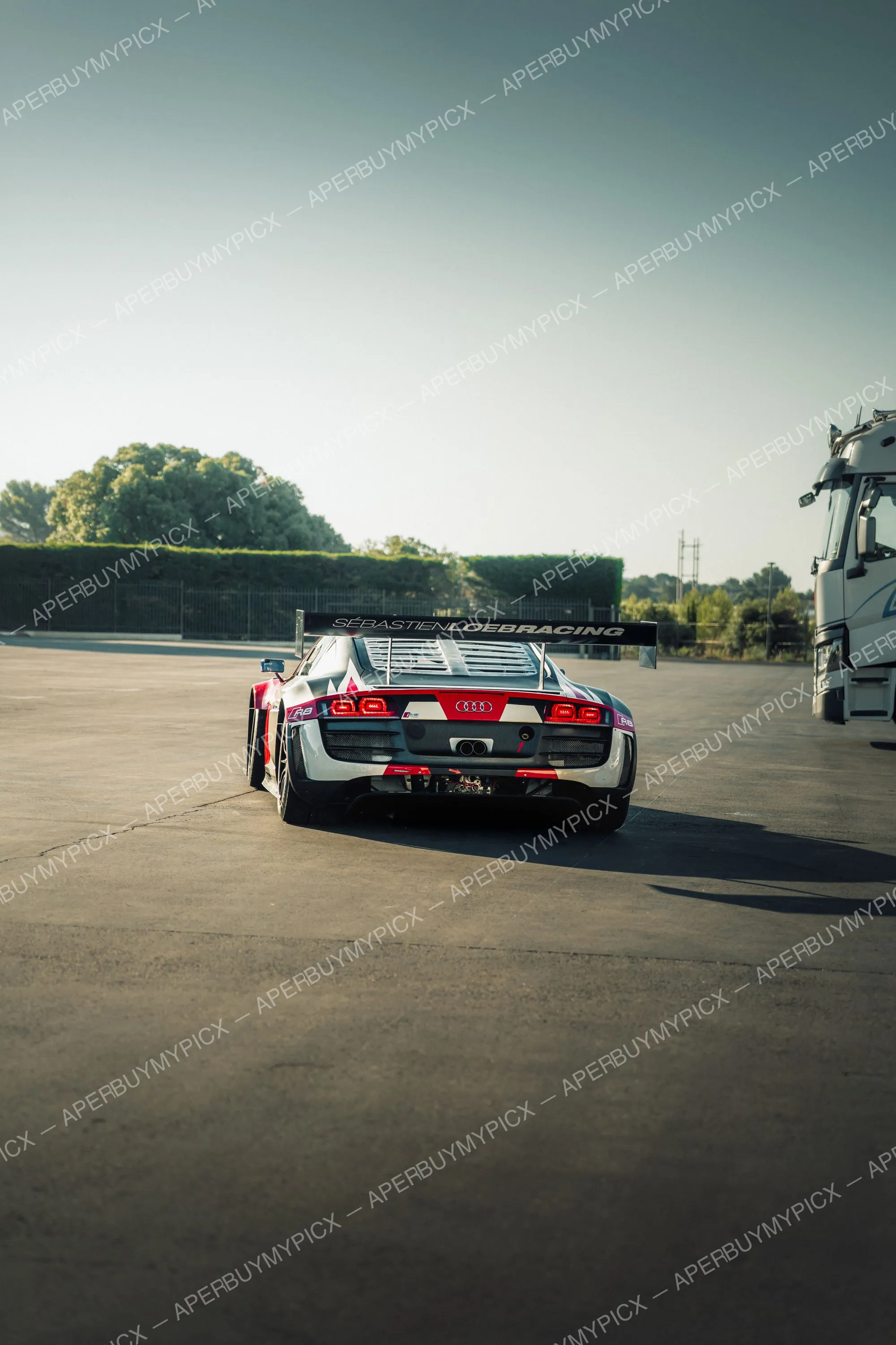 Vue arrière d'une Audi R8 LMS GT3 aux couleurs Sébastien Loeb Racing, gris, rouge et blanc, avec aileron arrière, stationnée sur un paddock ensoleillé près d'un camion, arbres et ciel dégagé en fond.