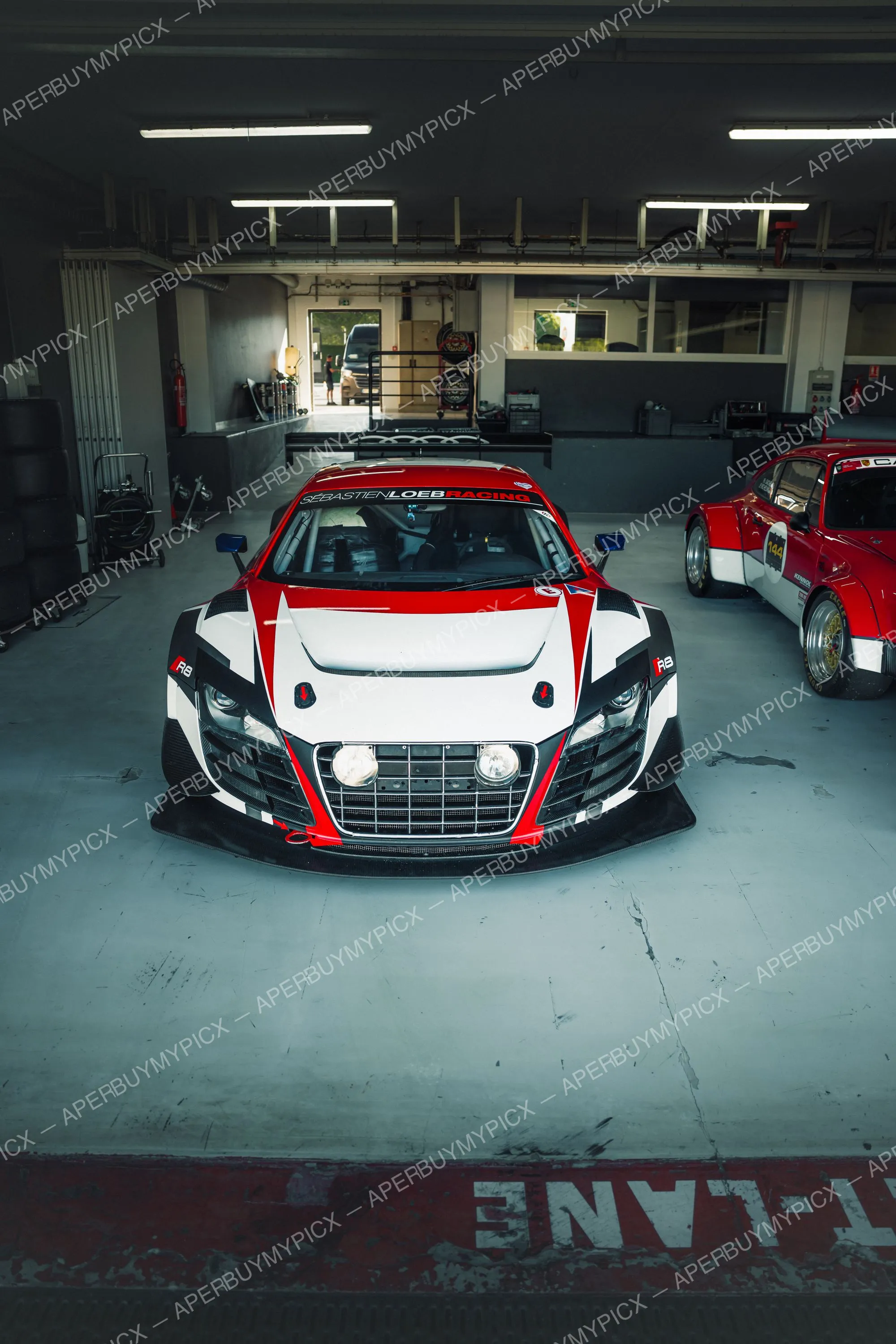 Audi R8 LMS GT3 blanche et rouge aux couleurs Sébastien Loeb Racing garée de face dans un stand de circuit, avec une Porsche 911 rouge n°144 à droite et l'inscription pit lane au sol.