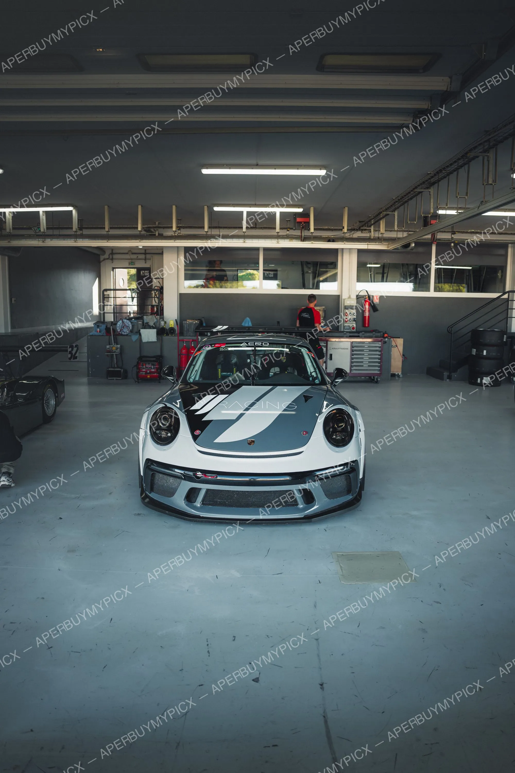 Porsche 911 GT3 Cup grise et noire aux couleurs Racing garée de face dans un box de circuit, avec mécanicien, caisses à outils et pneus en arrière-plan.