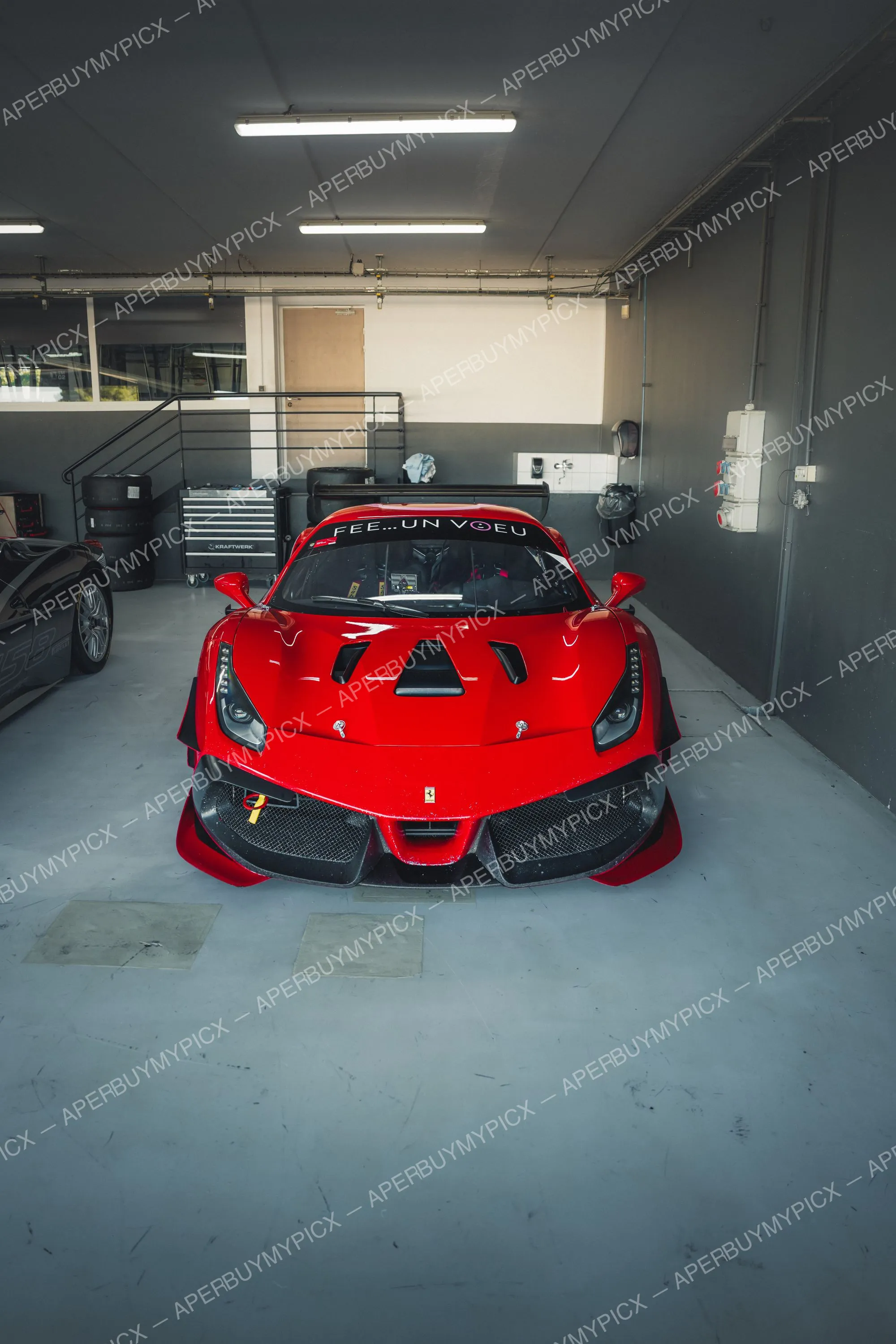 Ferrari 488 Challenge rouge vue de face, garée dans un box de garage au circuit du Castellet, avec une seconde voiture noire et des pneus racing à gauche.