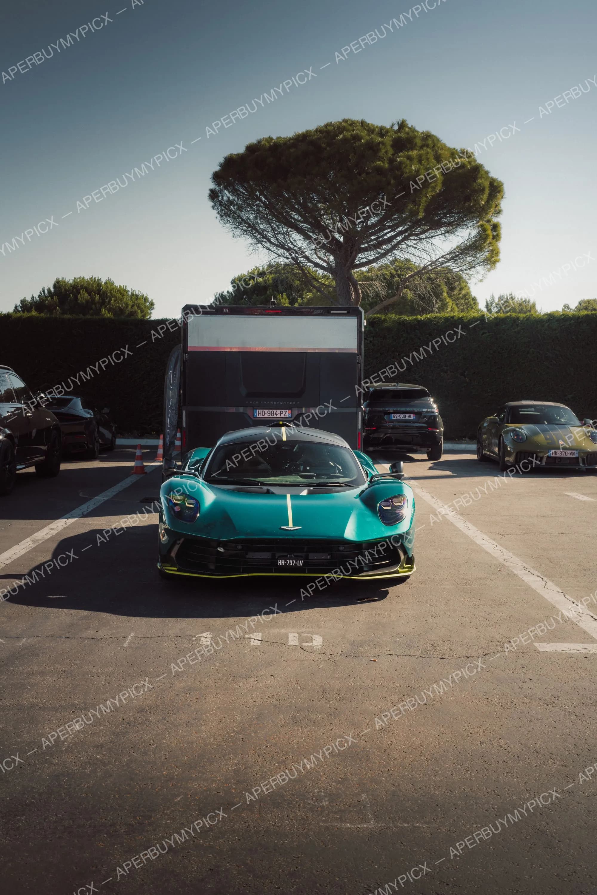 Aston Martin Valhalla verte avec bande jaune vue de face, garée sur un parking devant un camion de transport et un pin parasol, avec un Porsche 911 et un Range Rover à proximité.