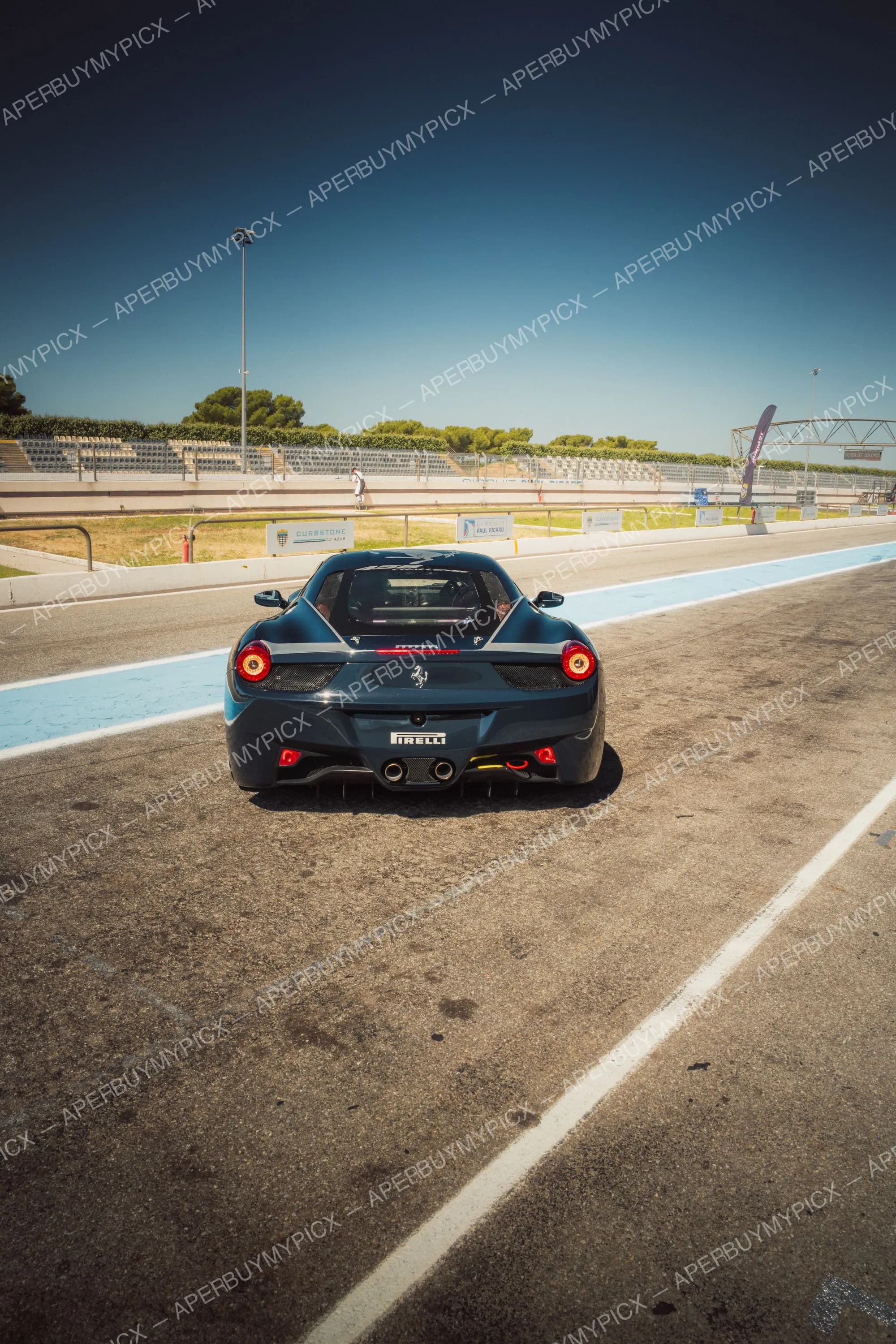 Vue arrière d'une Ferrari 458 bleu foncé avec bande grise et logo Pirelli, à l'arrêt sur la voie des stands du circuit Paul Ricard au Castellet, sous un ciel bleu dégagé.