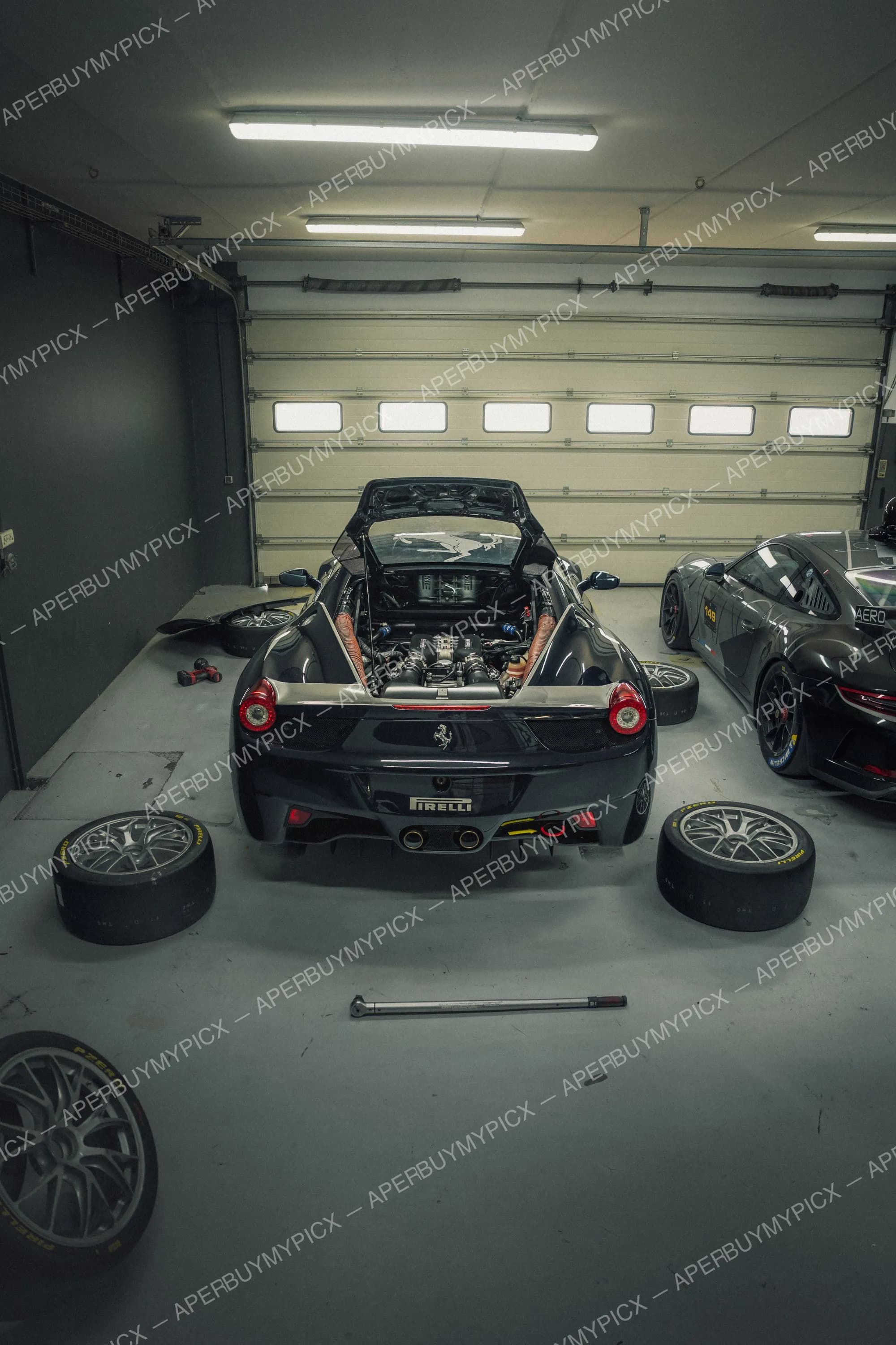 Ferrari 458 noire avec capot moteur arrière ouvert révélant le V8, garée dans un garage avec des pneus de course Pirelli et une clé dynamométrique au sol, une Porsche de course à côté.