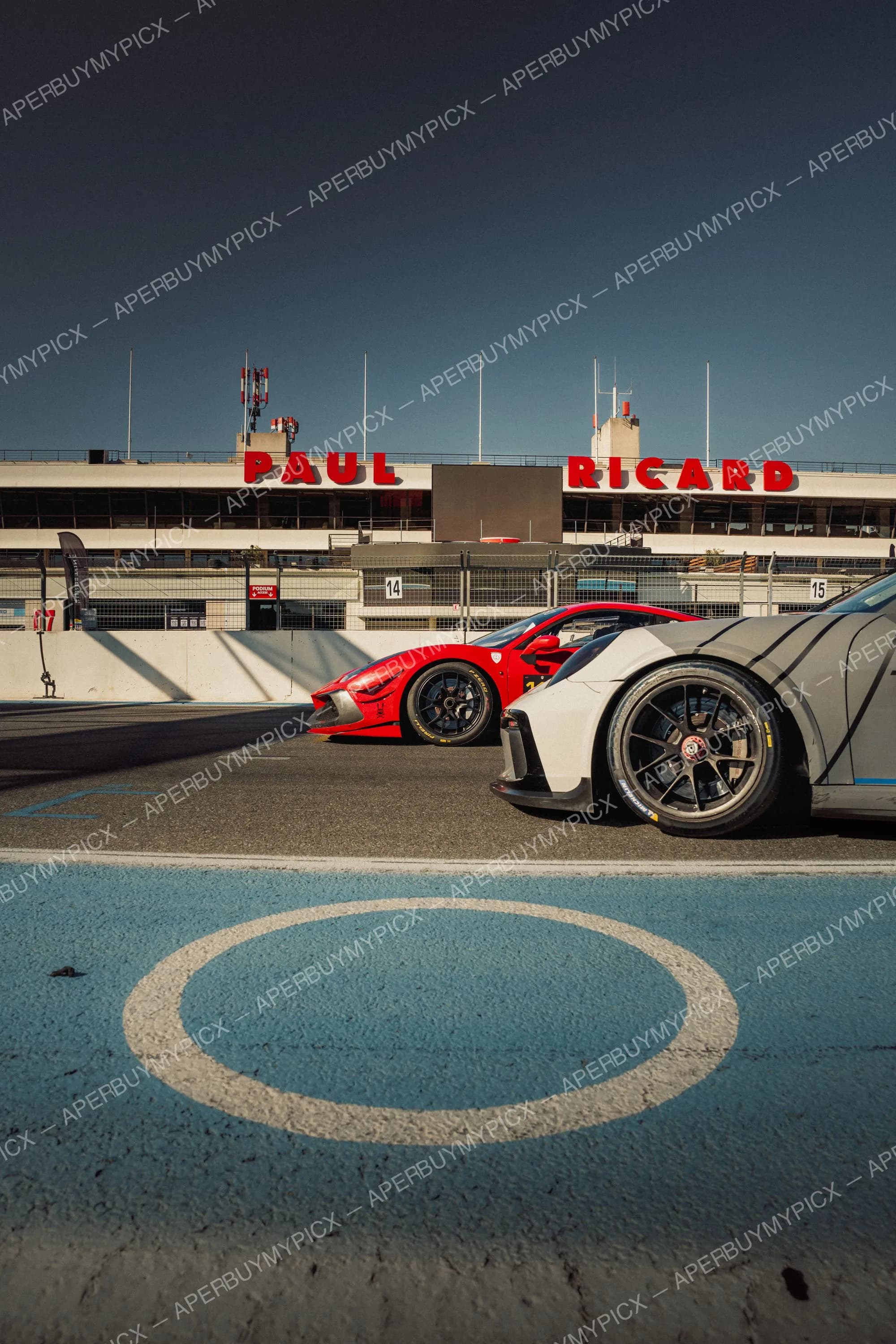 Une Ferrari GT rouge et une Porsche GT blanche stationnées dans la voie des stands du circuit Paul Ricard, avec l'enseigne rouge PAUL RICARD sur le bâtiment en arrière-plan et un ciel bleu clair.