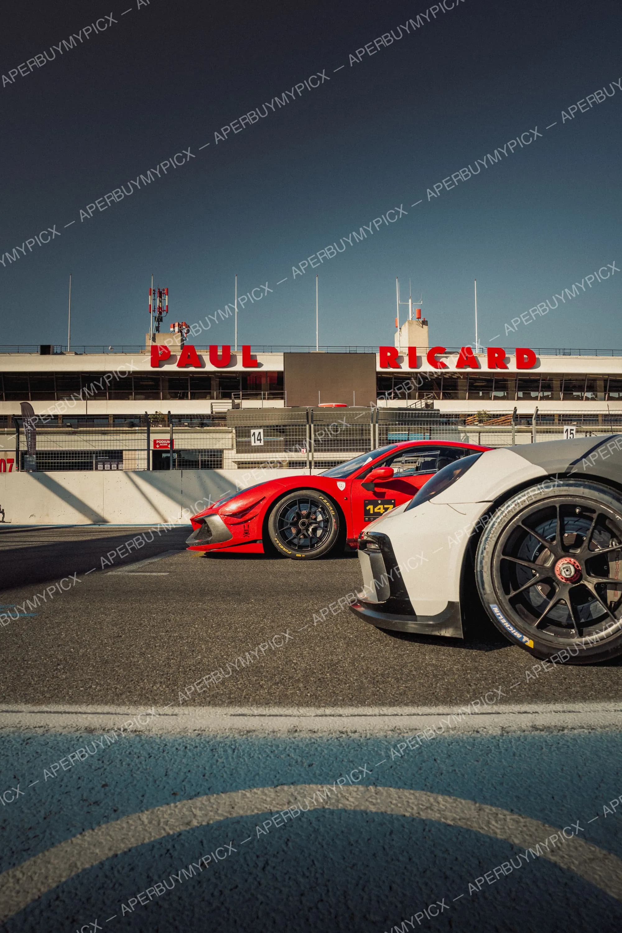 Ferrari 488 GT rouge numéro 147 sur pneus Pirelli et voiture blanche sur pneus Michelin sur la piste du circuit Paul Ricard, devant les tribunes avec l'enseigne rouge PAUL RICARD sous un ciel bleu.
