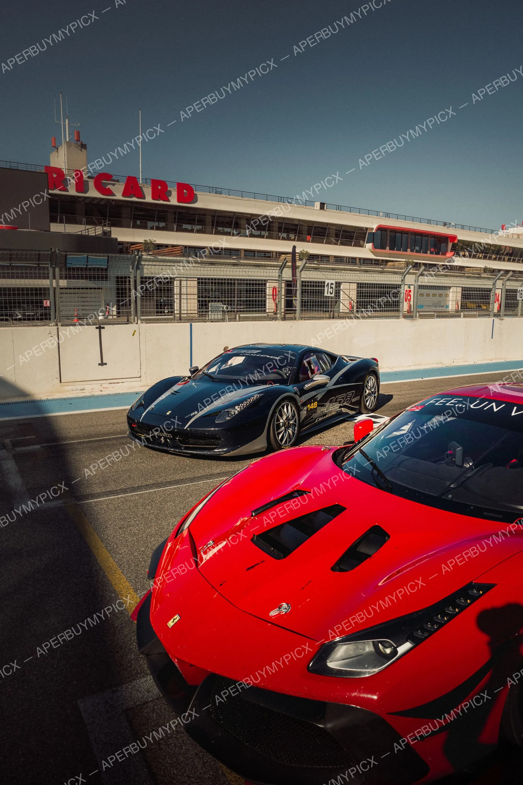 Ferrari 458 Challenge noire et Ferrari rouge dans la voie des stands du circuit Paul Ricard, sous la tribune RICARD, ciel bleu.