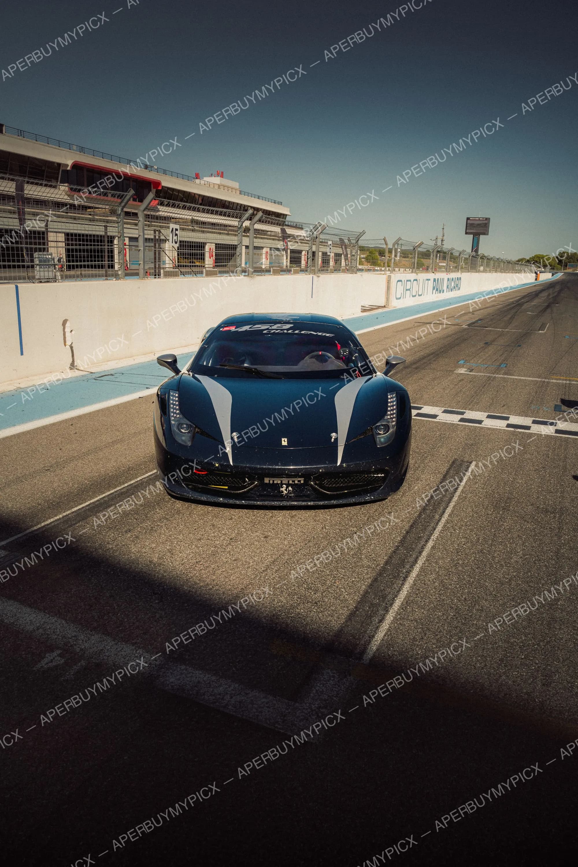 Ferrari 458 Challenge bleu foncé vue de face dans la pit lane du Circuit Paul Ricard, sous un ciel dégagé, avec les stands et la ligne d'arrivée à damier.