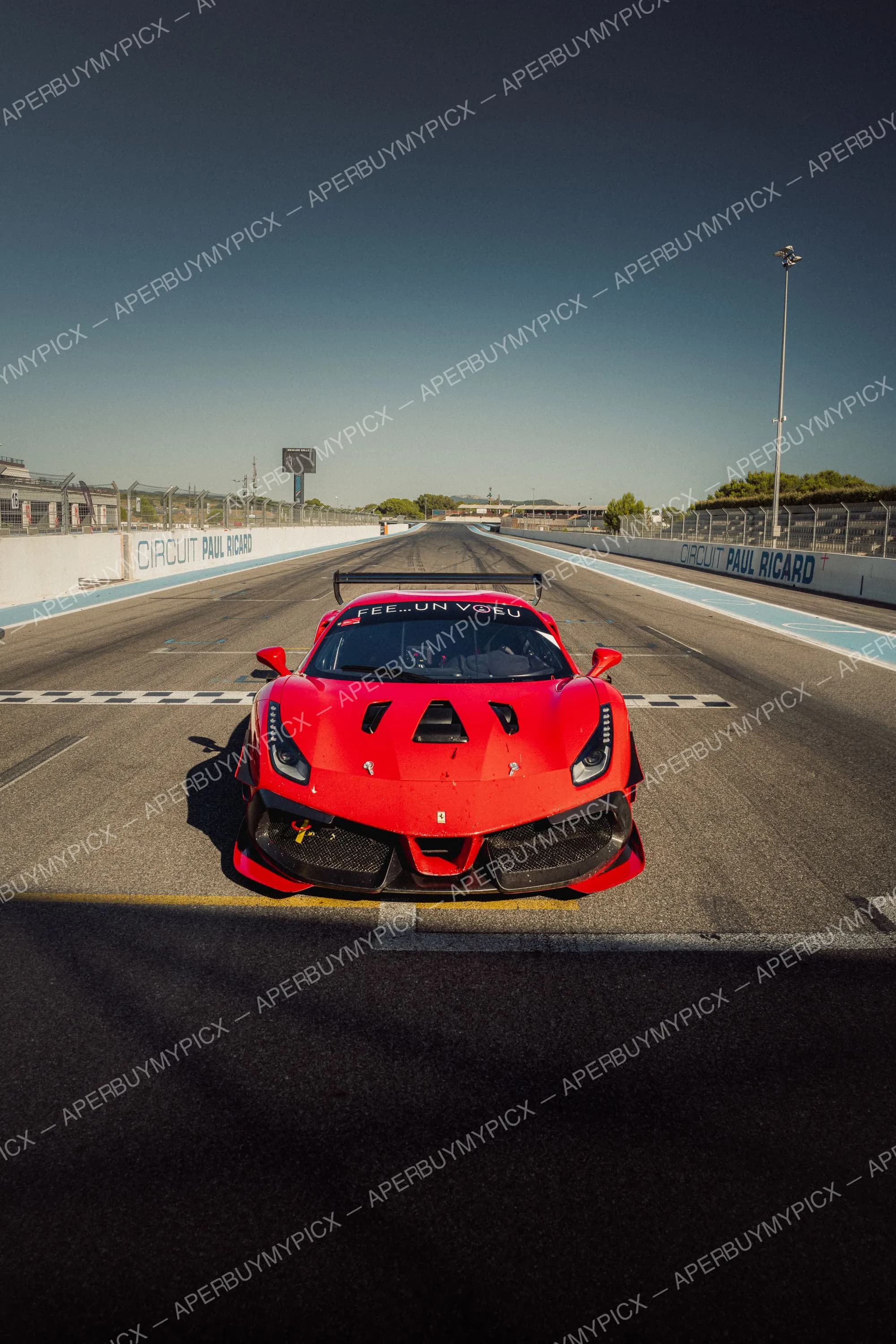 Ferrari 488 GT3 rouge de face sur la ligne de départ du Circuit Paul Ricard, sous un ciel bleu clair, avec les murets bleus et blancs de la piste.