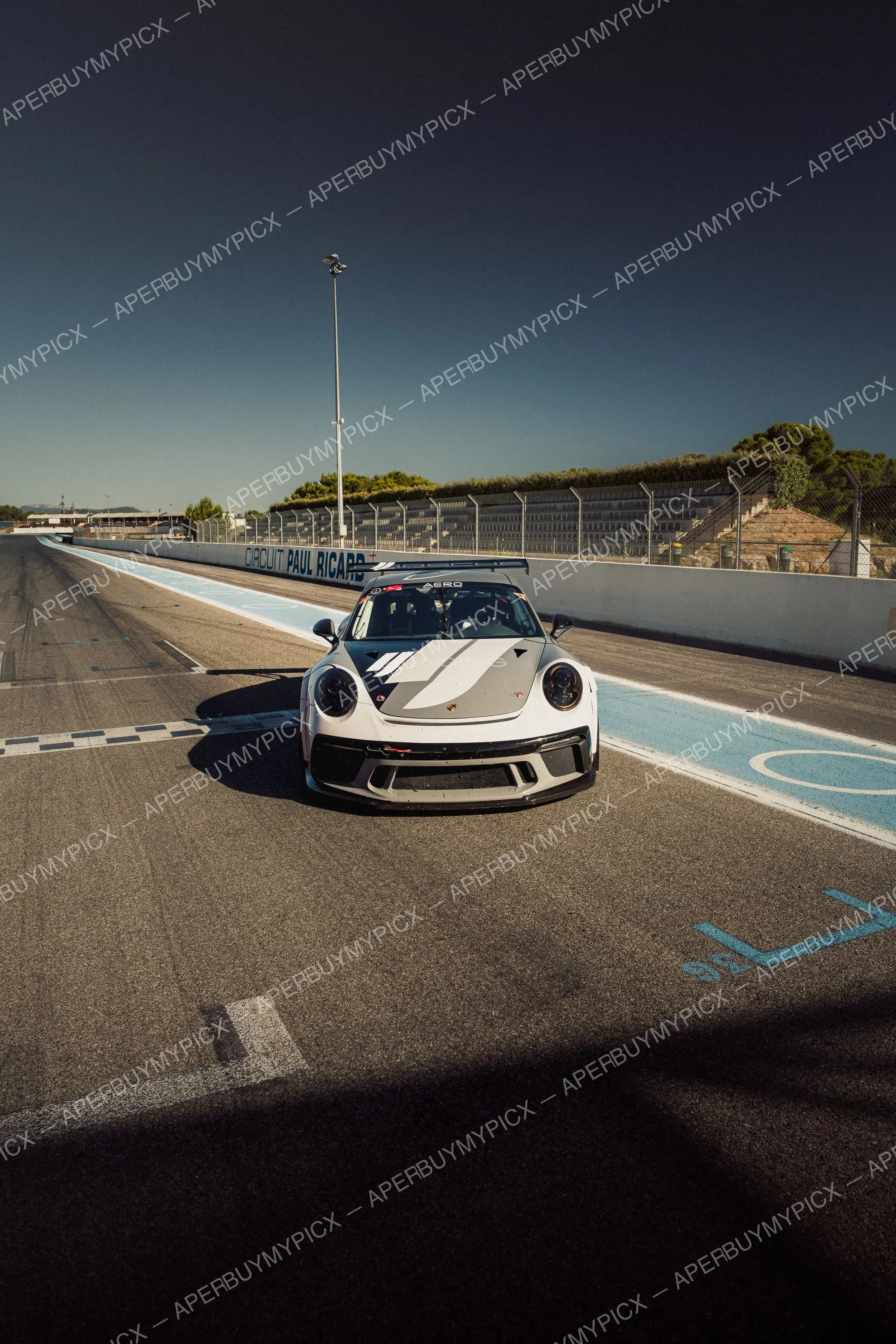 Porsche 911 GT3 Cup blanche vue de face dans la voie des stands du Circuit Paul Ricard, sous un ciel bleu dégagé avec tribunes et clôtures en arrière-plan.