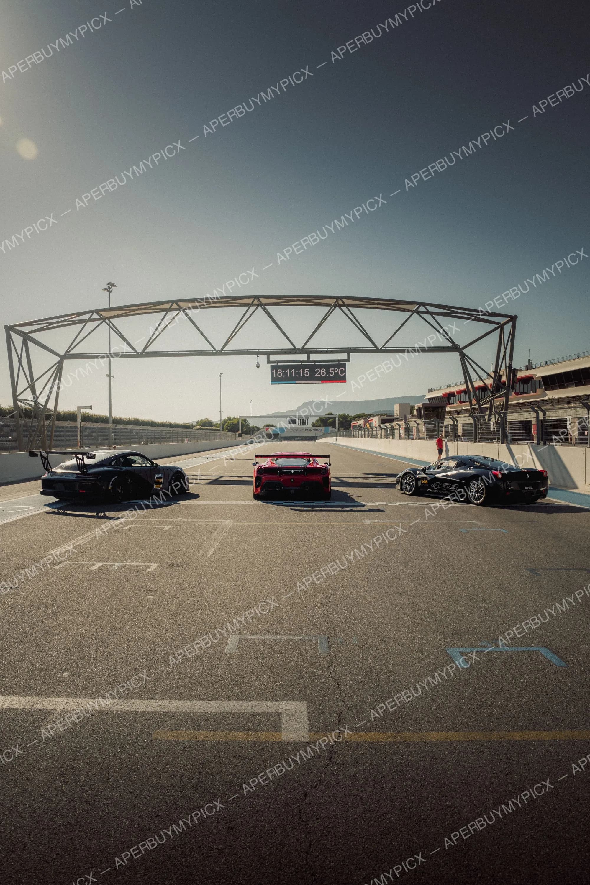 Trois voitures de course Ferrari, deux noires et une rouge, alignées sur la piste du Circuit Paul Ricard sous un ciel dégagé, panneau affichant 18:11:15 et 26,5°C.