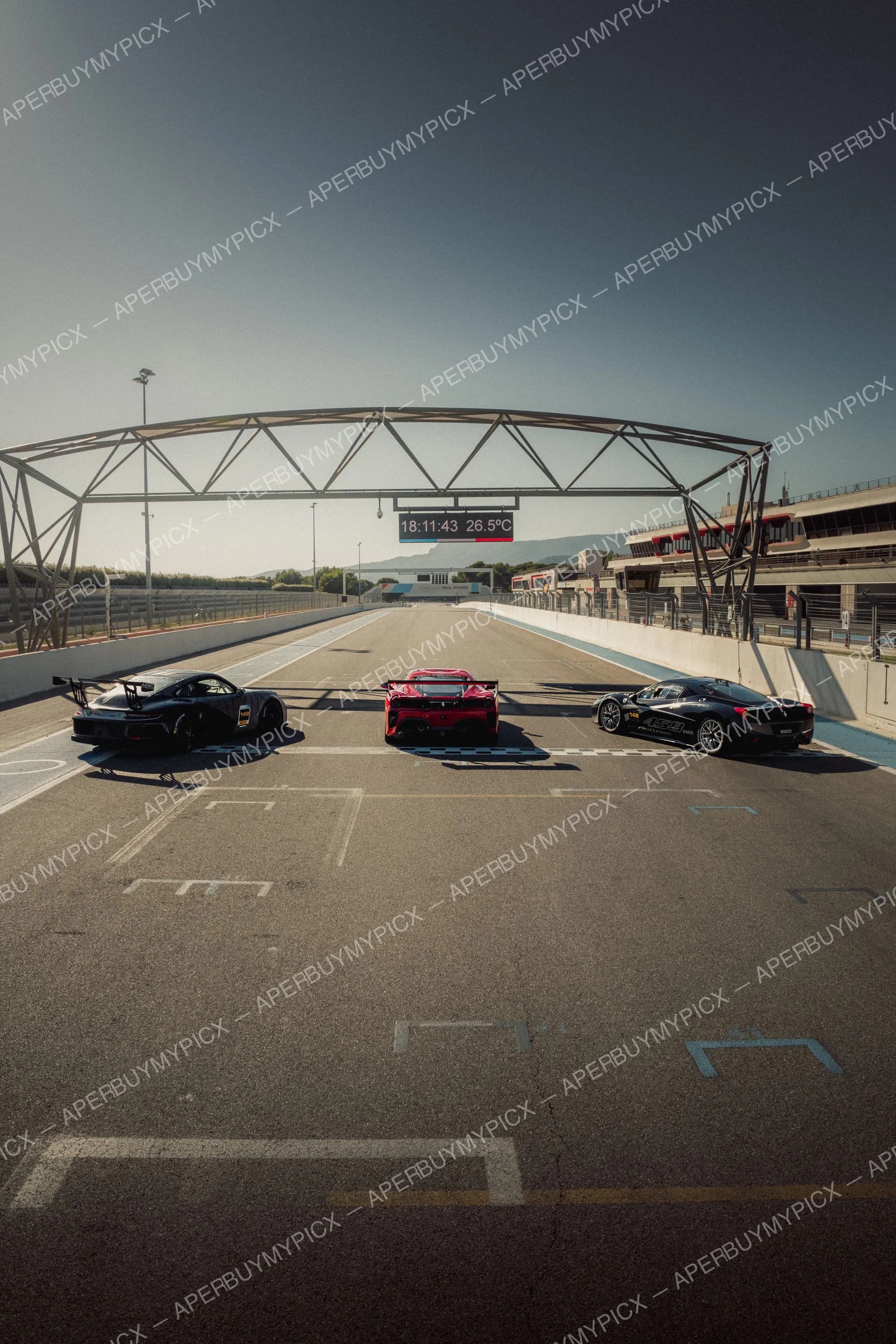 Trois voitures GT, une Porsche noire, une Ferrari 488 rouge et une Ferrari 458 Challenge noire, alignées sur la ligne de départ du Circuit Paul Ricard sous un ciel dégagé, avec le portique d'arrivée et l'affichage 18:11:43 26,5°C.