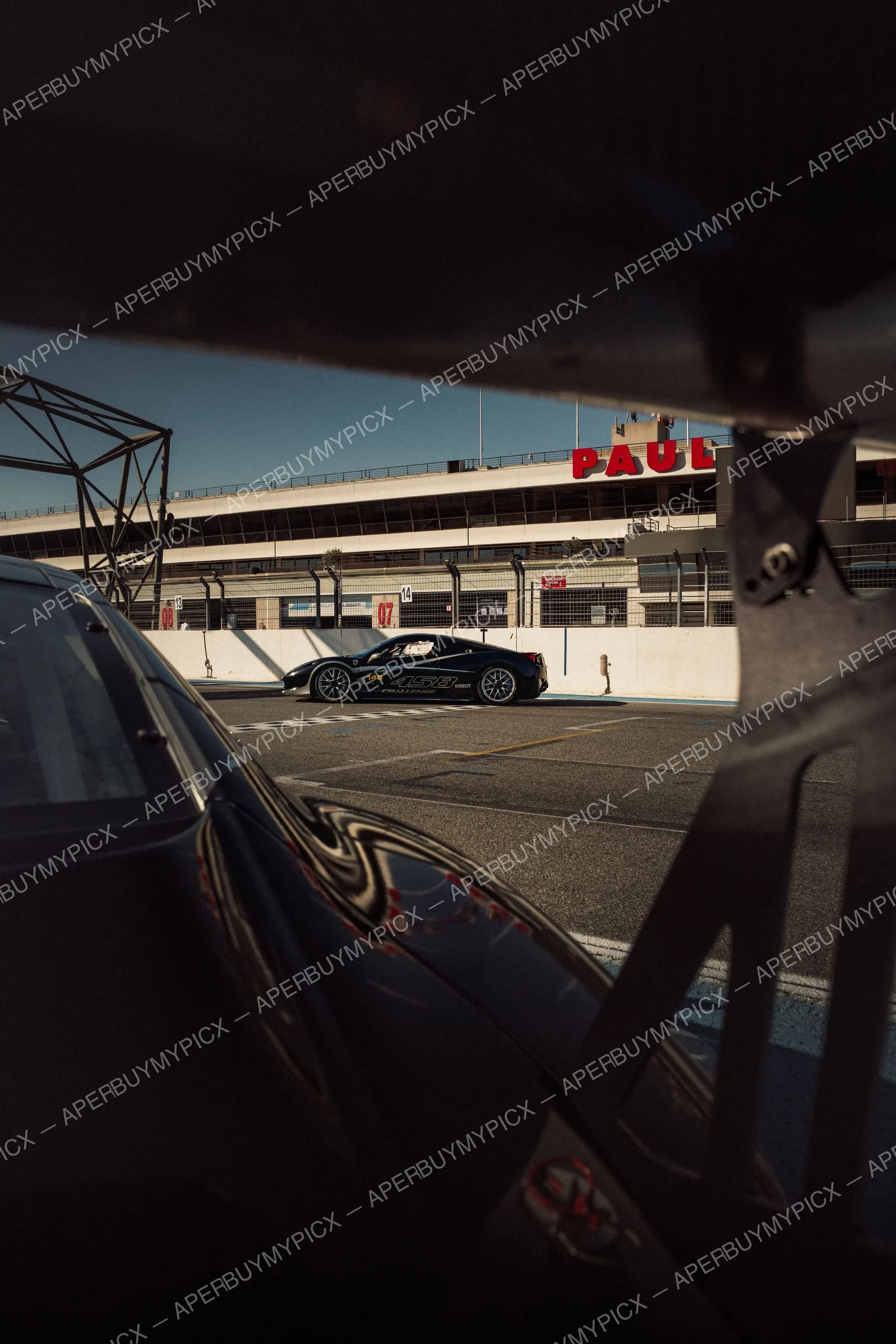Ferrari 458 Challenge noire dans la voie des stands du Circuit Paul Ricard, vue à travers l'aileron et la carrosserie d'une autre voiture, avec les tribunes en arrière-plan.