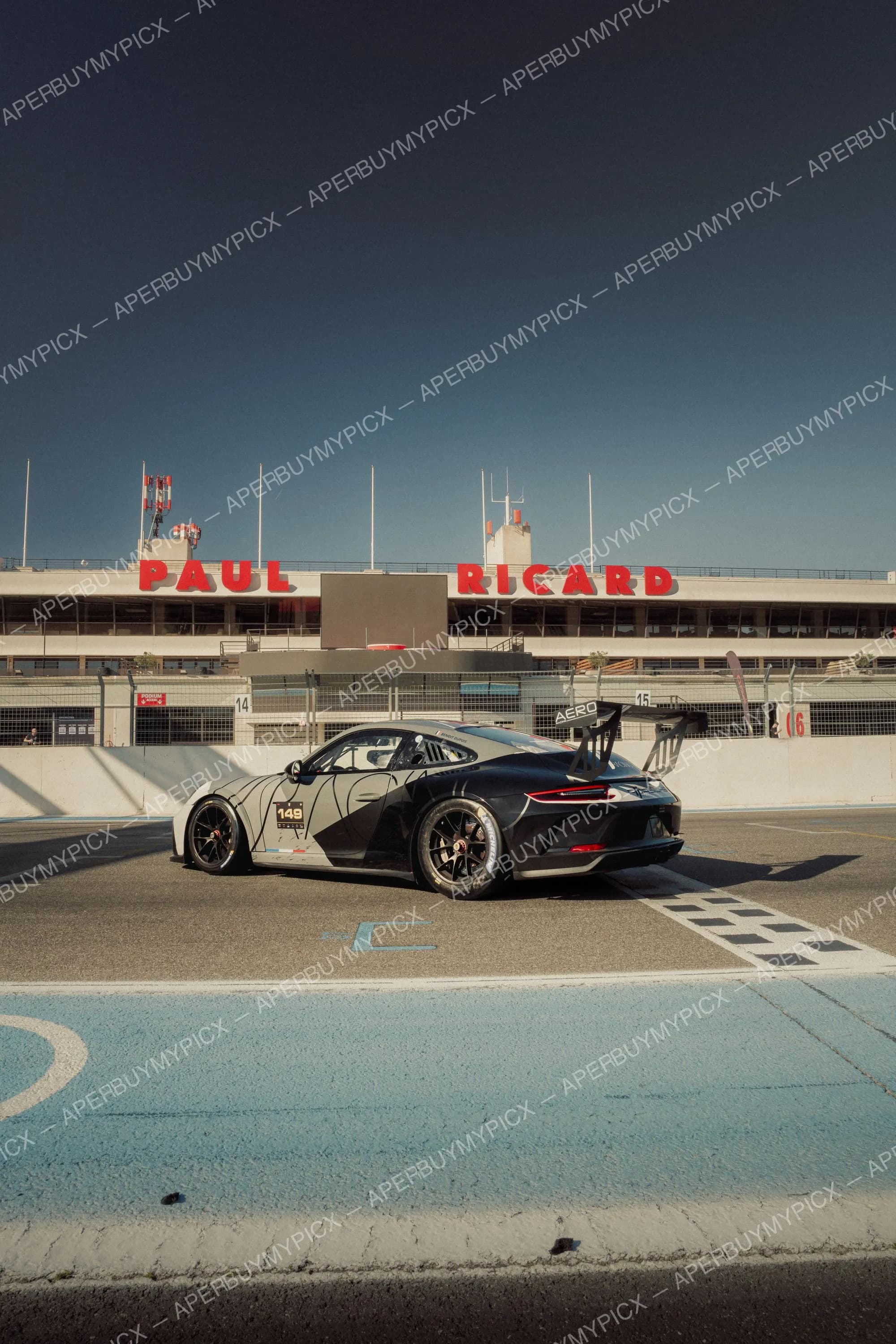 Porsche 911 GT3 Cup noire et grise numéro 149 stationnée sur la voie des stands du Circuit Paul Ricard, avec la tribune et l'enseigne rouge PAUL RICARD en arrière-plan sous un ciel bleu.