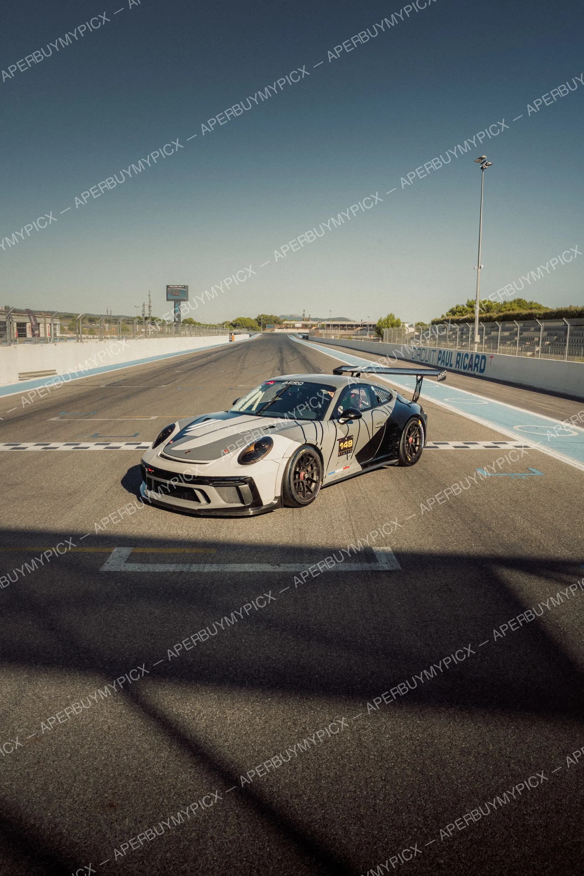Porsche 911 GT3 Cup grise et noire numéro 149 avec aileron arrière, arrêtée sur la ligne de départ du Circuit Paul Ricard sous un ciel bleu dégagé.