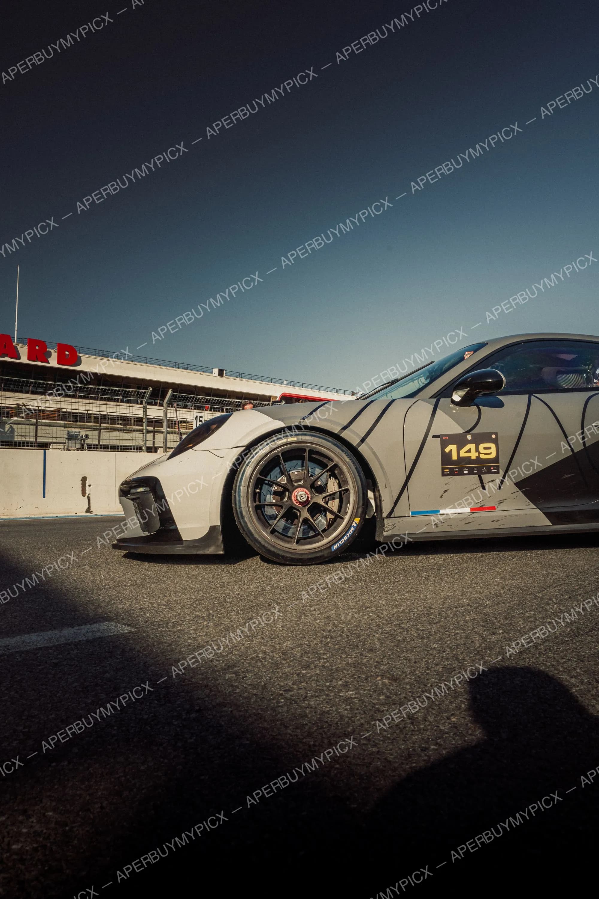 Porsche 911 GT3 grise numéro 149 avec jantes noires et pneus Michelin sur la piste du circuit Paul Ricard au Castellet, tribune en arrière-plan sous un ciel dégagé