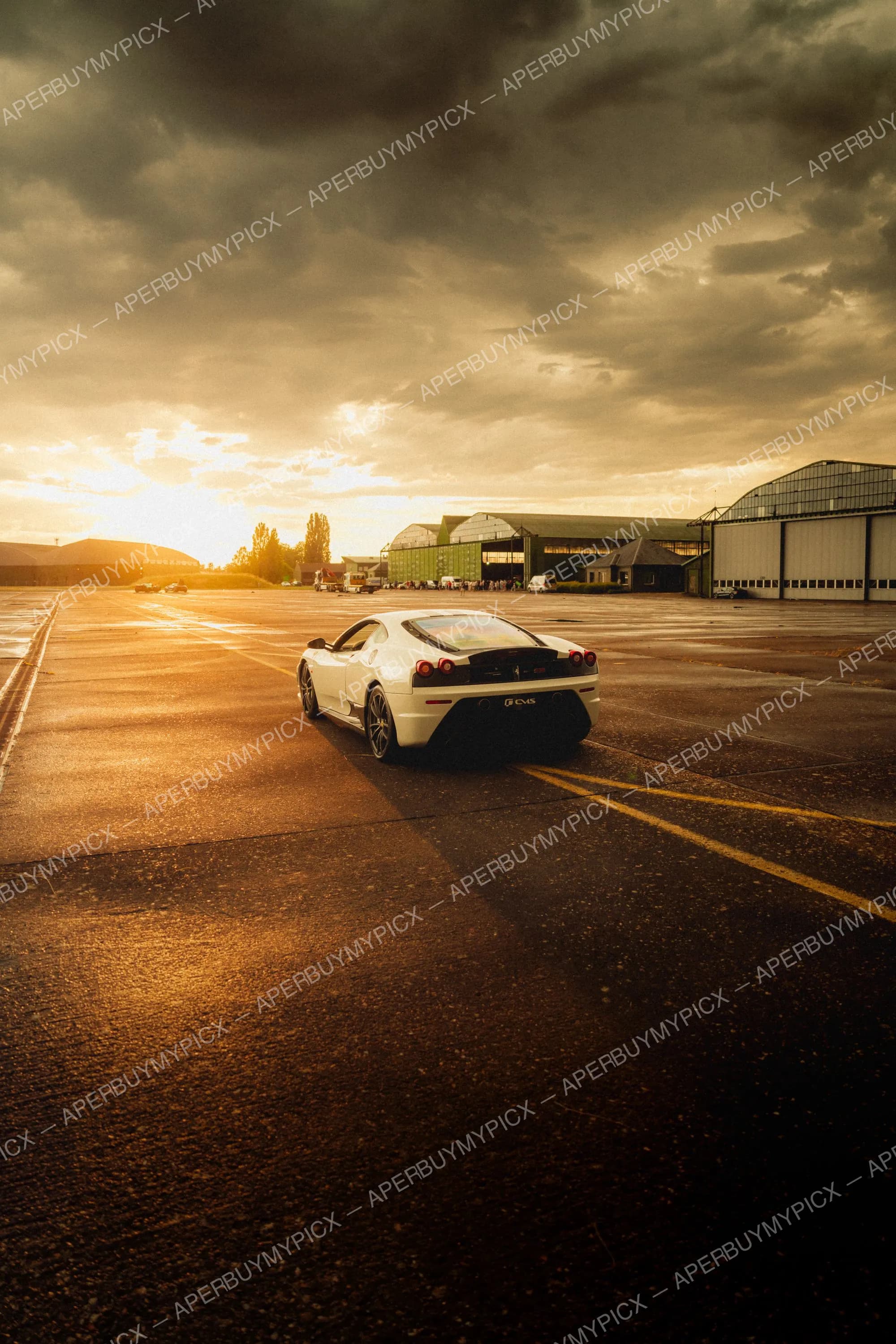 Ferrari F430 Scuderia blanche vue de trois quarts arrière sur un tarmac d'aérodrome au coucher de soleil, avec des hangars et un ciel nuageux doré en arrière-plan.