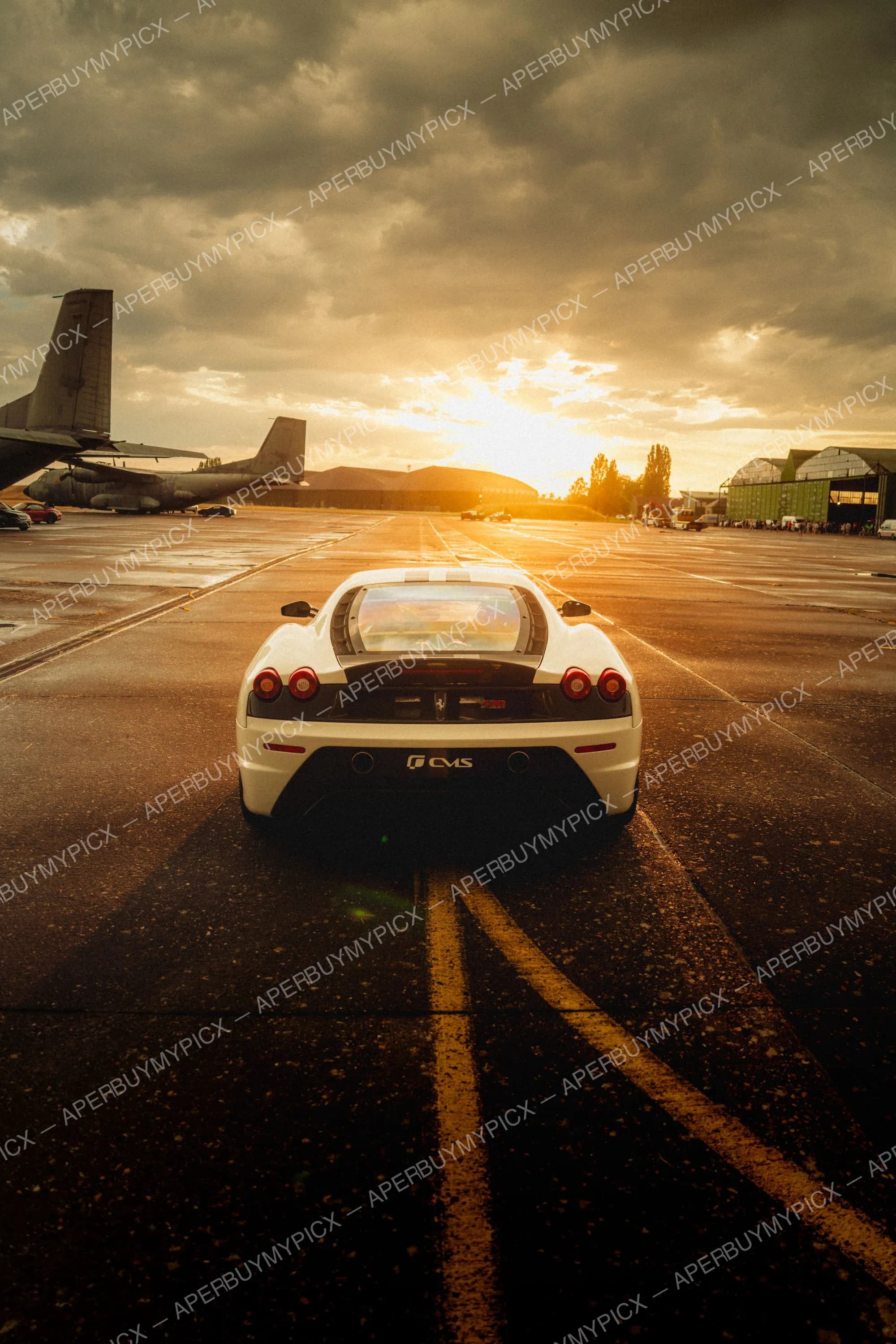 Ferrari 430 Scuderia blanche vue de l'arrière sur une piste d'aéroport au coucher du soleil, avec des avions militaires et des hangars en arrière-plan