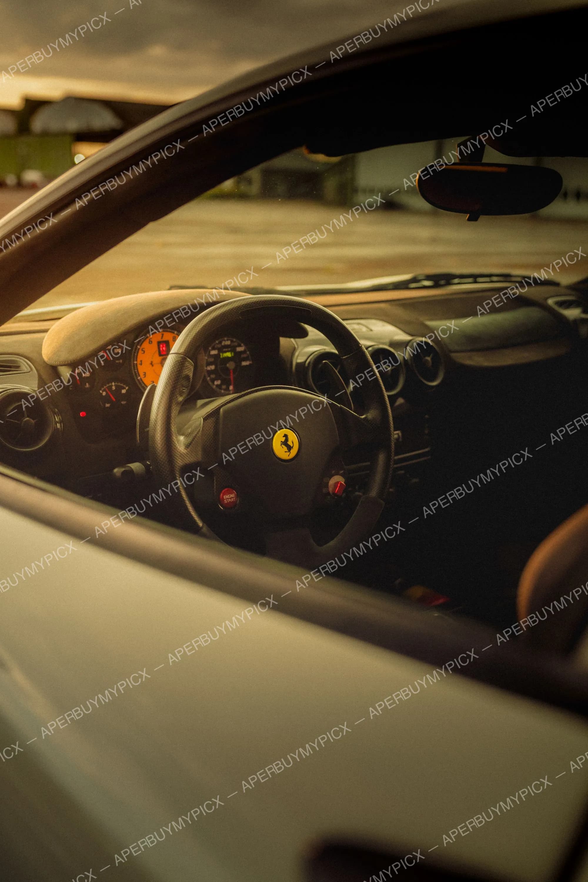 Vue de l'intérieur d'une Ferrari F430 à travers la vitre côté conducteur, montrant le volant en cuir avec logo cheval cabré jaune, le bouton Engine Start rouge et le tableau de bord avec compteurs oranges, au coucher du soleil.