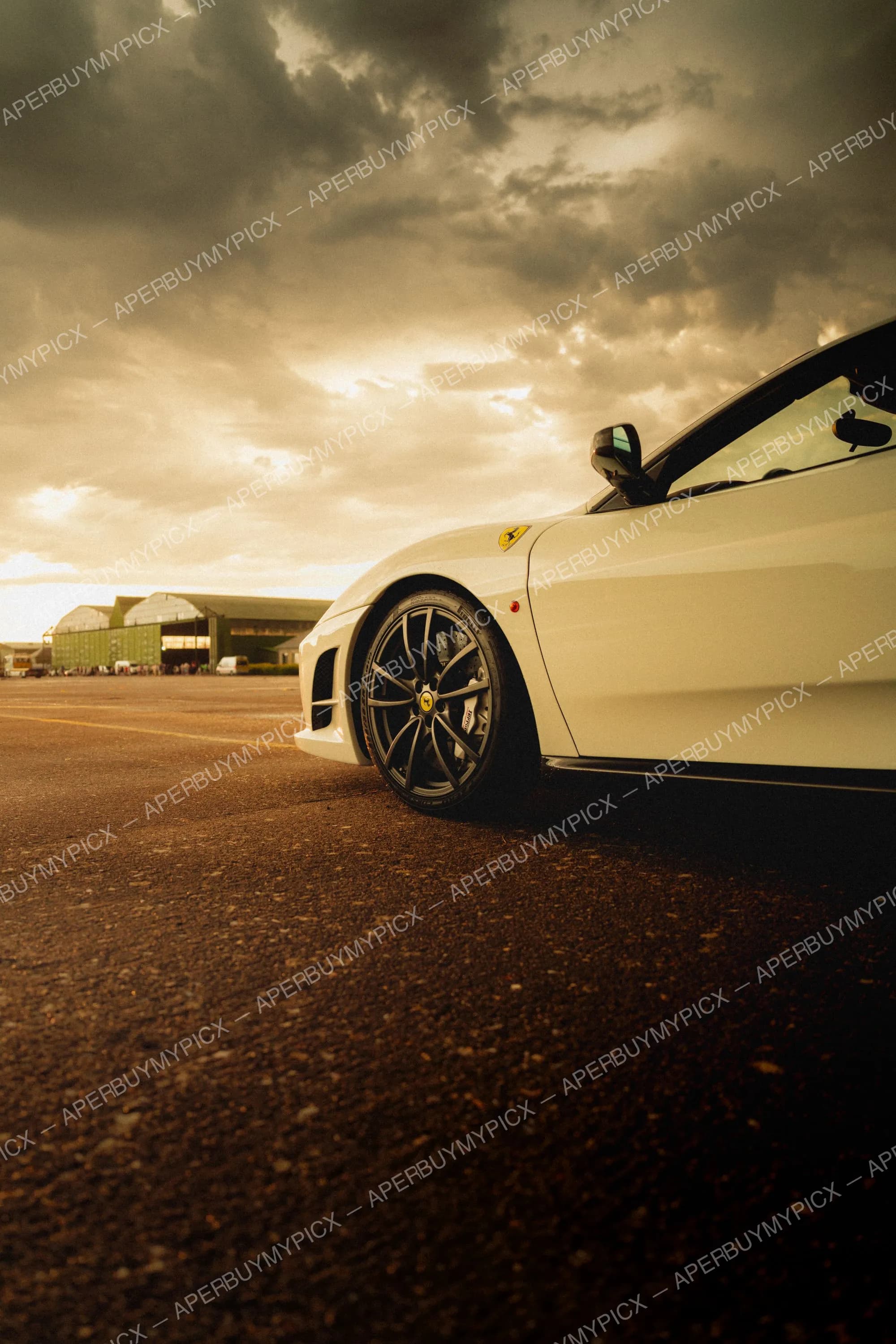 Ferrari F430 blanche vue en contre-plongée sur un tarmac d'aérodrome, avec jantes noires, étriers de frein rouges, sous un ciel nuageux au coucher de soleil et des hangars verts en arrière-plan.