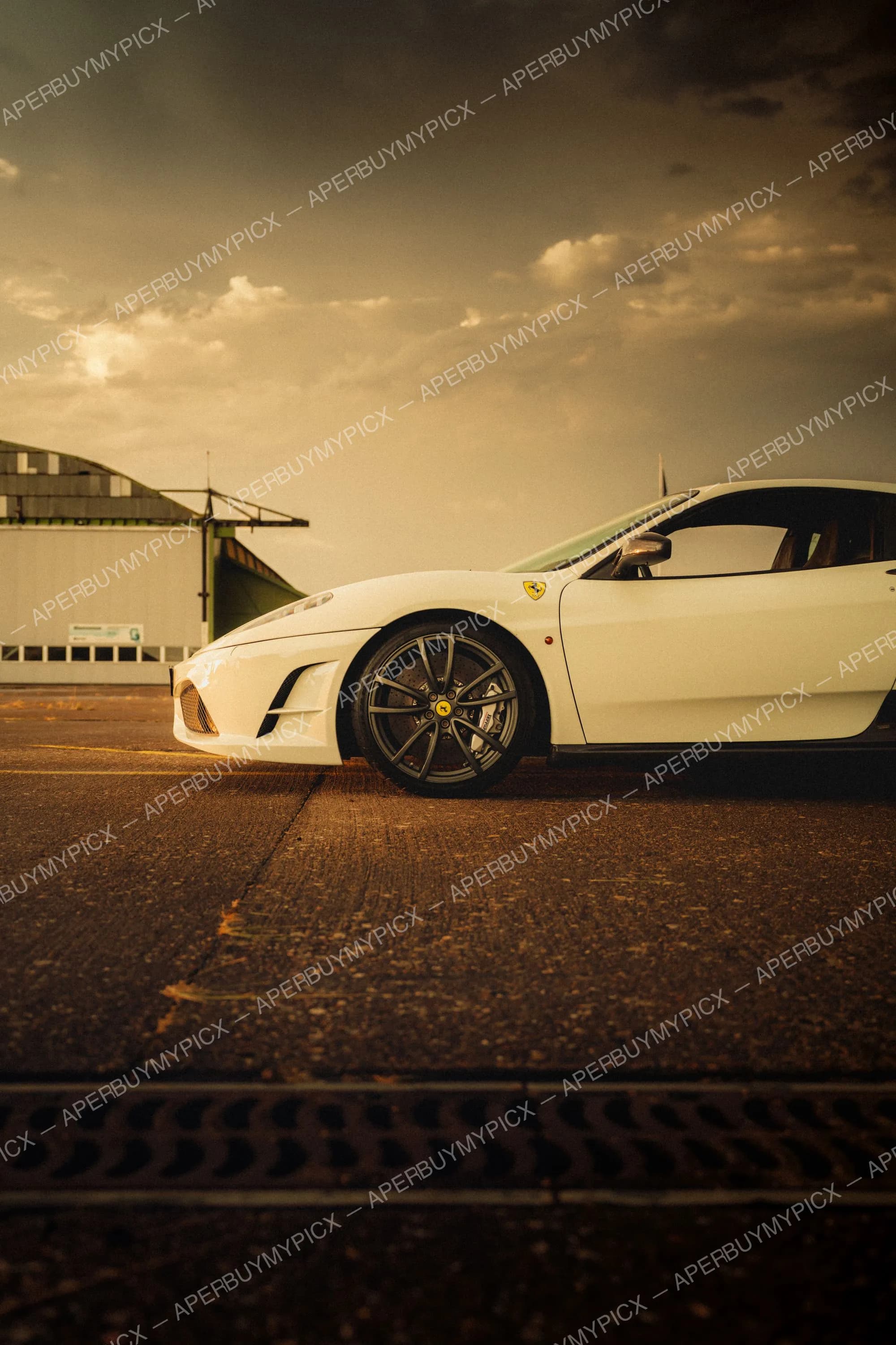 Ferrari F430 Scuderia blanche vue de profil garée sur une piste au coucher de soleil, jantes noires et étriers de frein jaunes, hangar en arrière-plan sous un ciel nuageux doré.