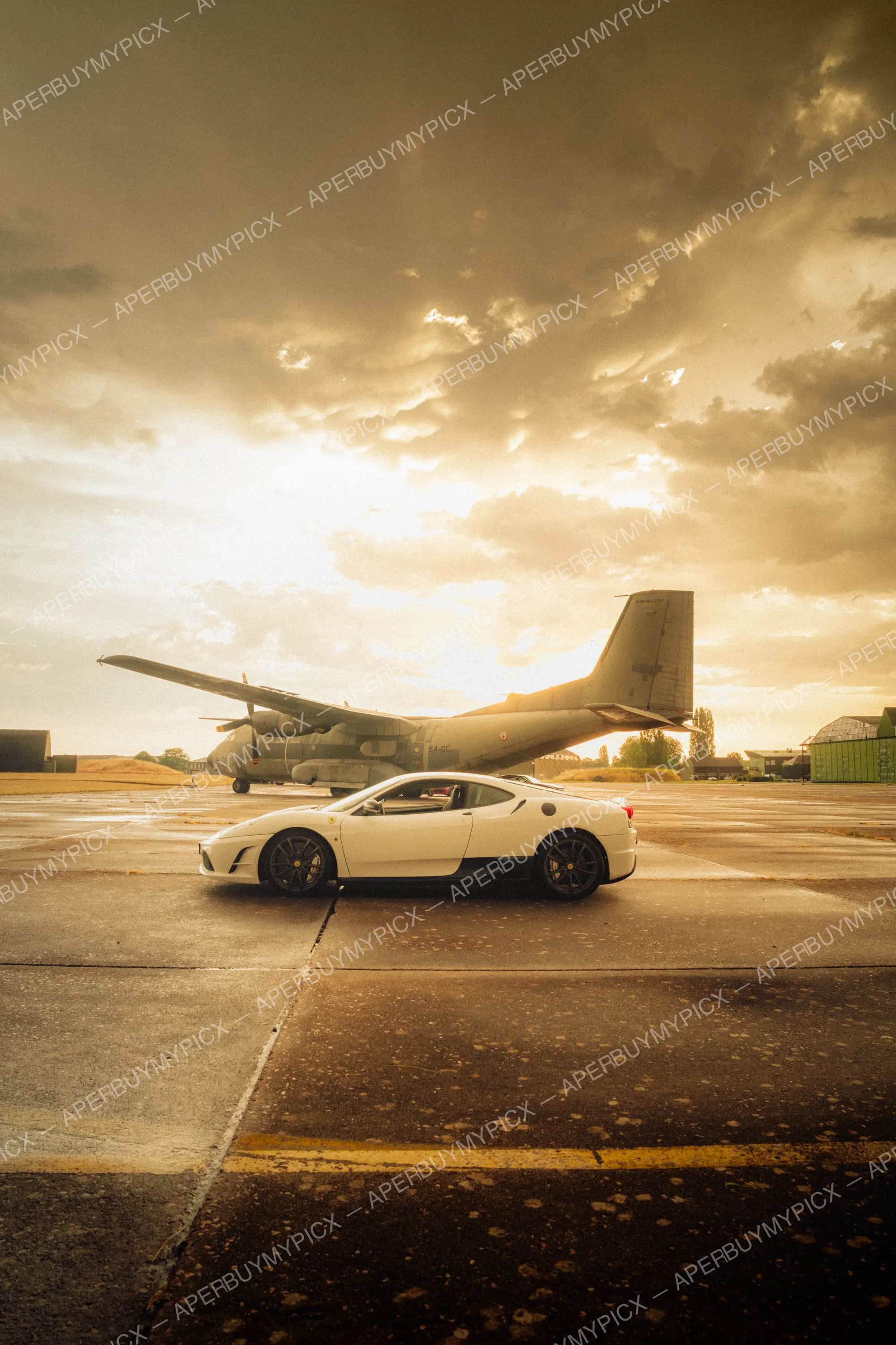 Ferrari F430 blanche vue de profil sur une piste d'aéroport, devant un avion de transport militaire Transall, sous un ciel nuageux doré au coucher du soleil
