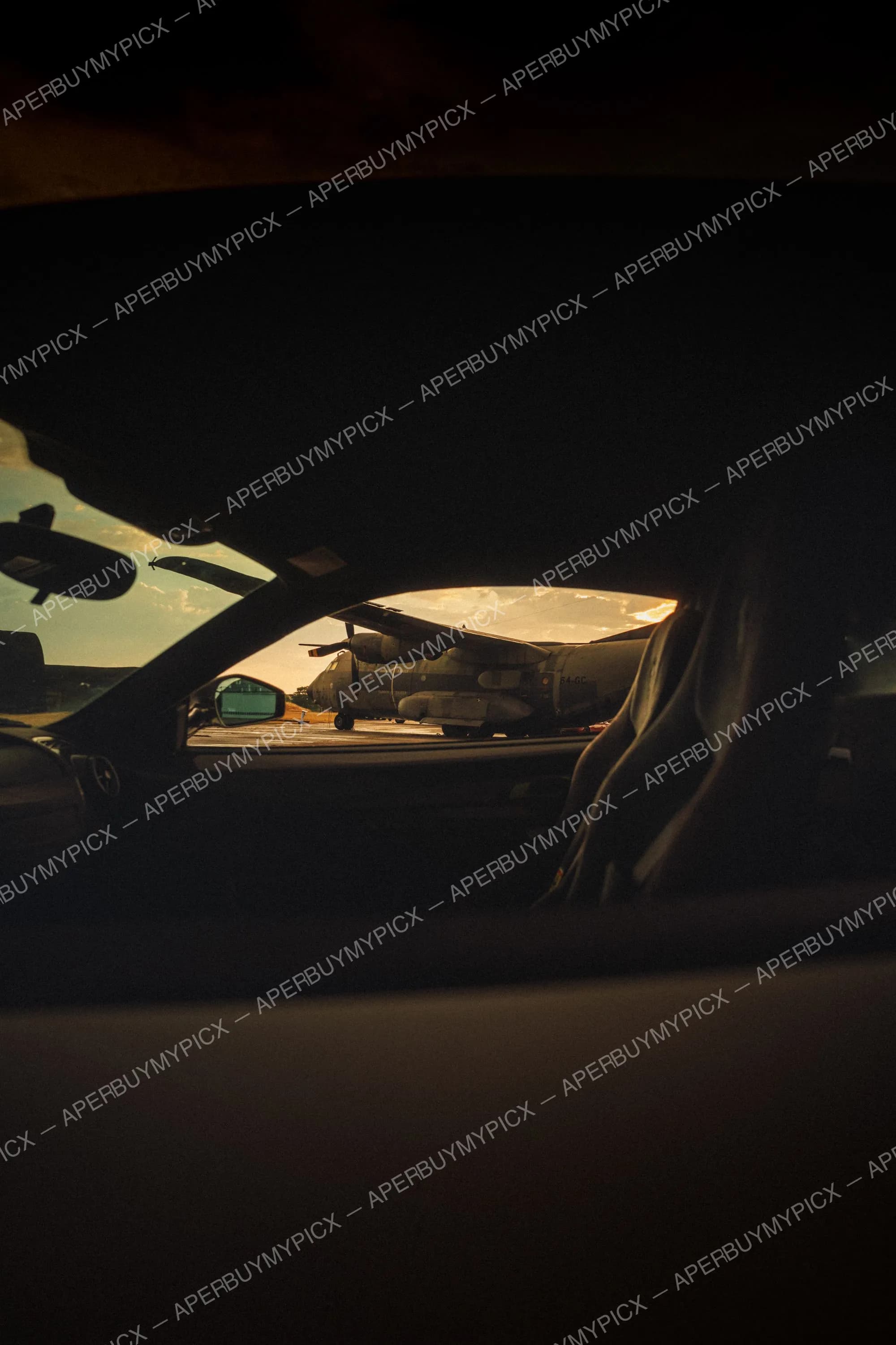 Vue depuis l'intérieur sombre d'un véhicule sur un avion de transport militaire stationné sur le tarmac, silhouette d'ailes et d'hélices se découpant sur un ciel orangé de coucher de soleil.