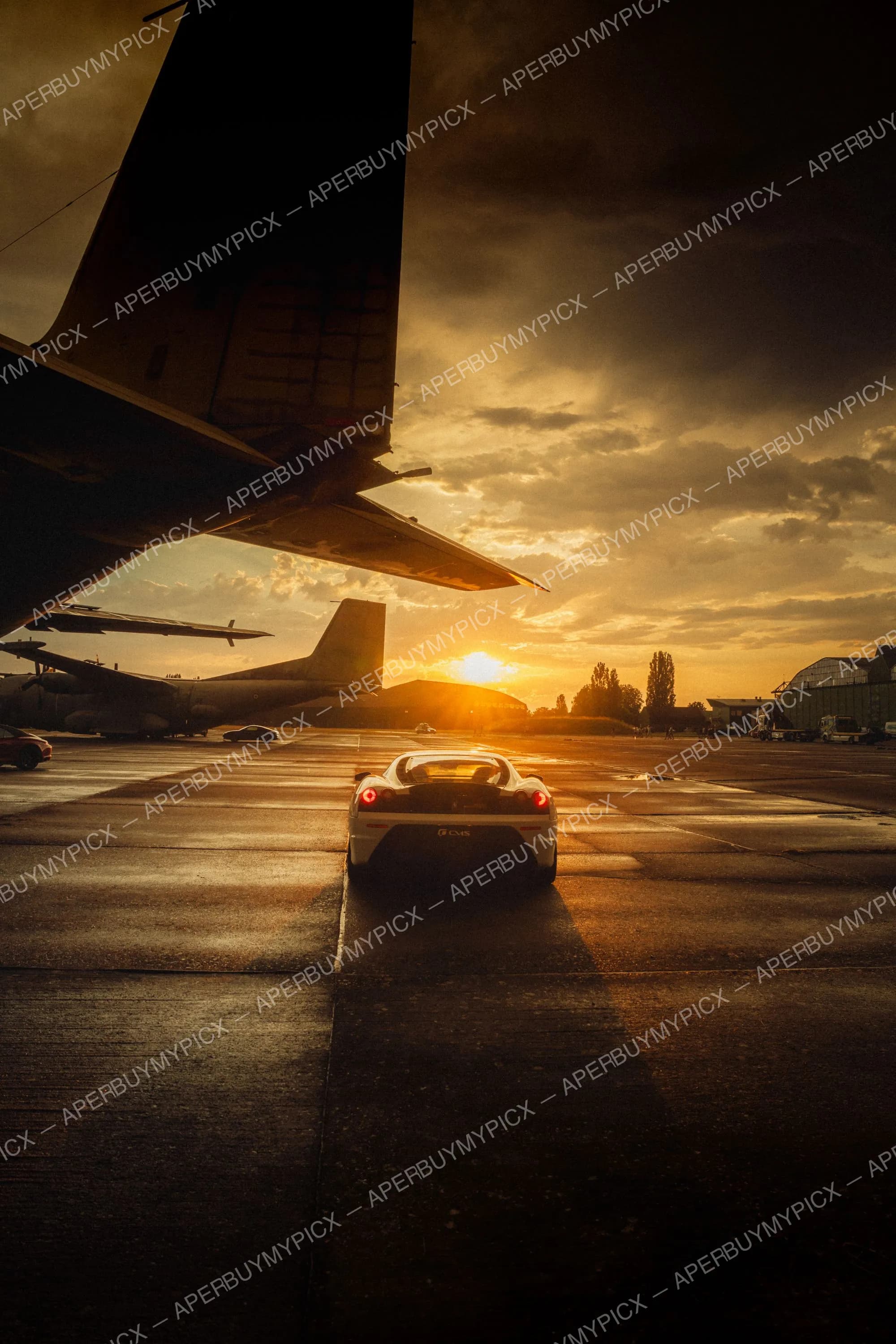 Supercar blanche vue de l'arrière sur le tarmac d'un aéroport au coucher de soleil, sous l'aile d'un avion, avec des avions vintage et des hangars en arrière-plan