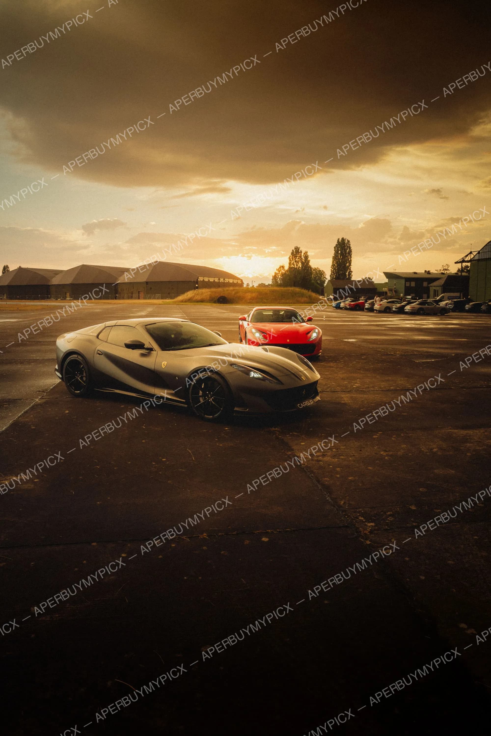 Deux Ferrari 812, une grise mate et une rouge, garées sur une piste d'aérodrome au coucher de soleil doré avec hangars et ciel nuageux en arrière-plan.