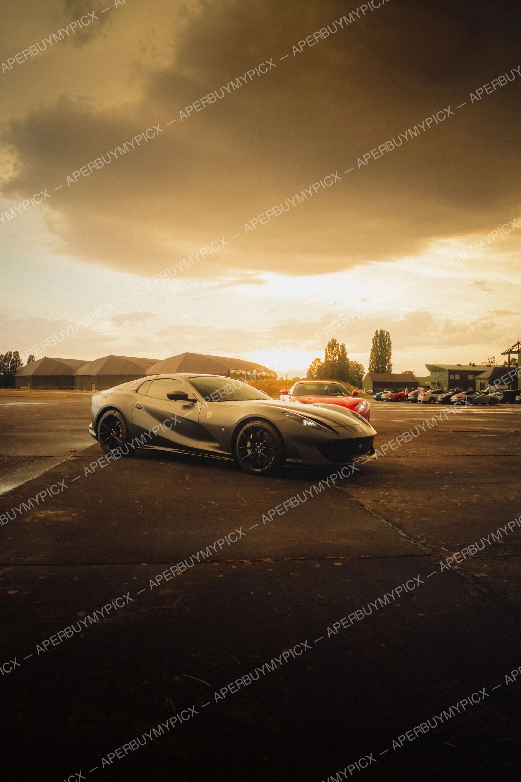 Ferrari 812 Superfast grise garée sur un tarmac au coucher de soleil, avec une Ferrari rouge et des hangars en arrière-plan sous un ciel nuageux doré.