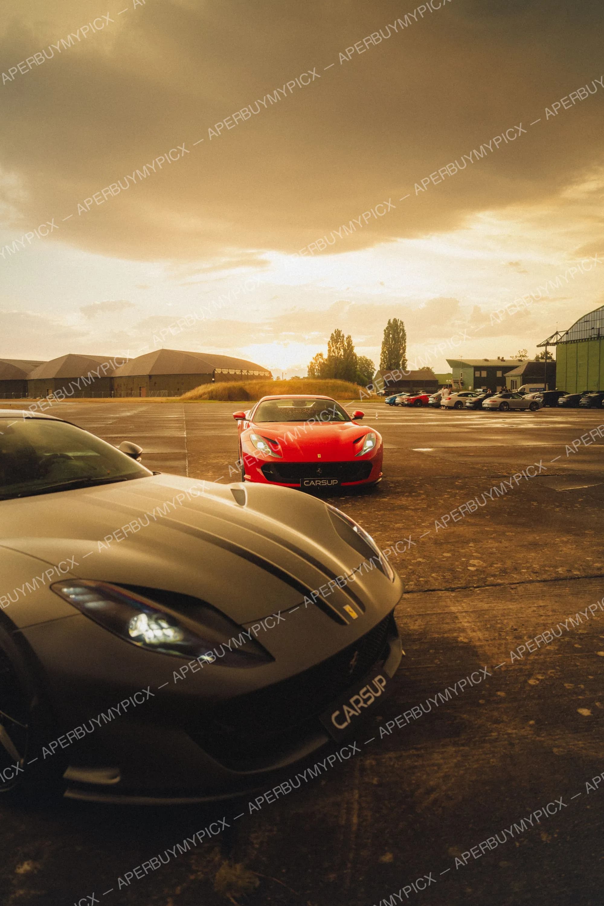 Deux Ferrari 812 Superfast, une rouge et une noire mate, garées sur un tarmac d'aérodrome au coucher de soleil doré avec hangars en arrière-plan