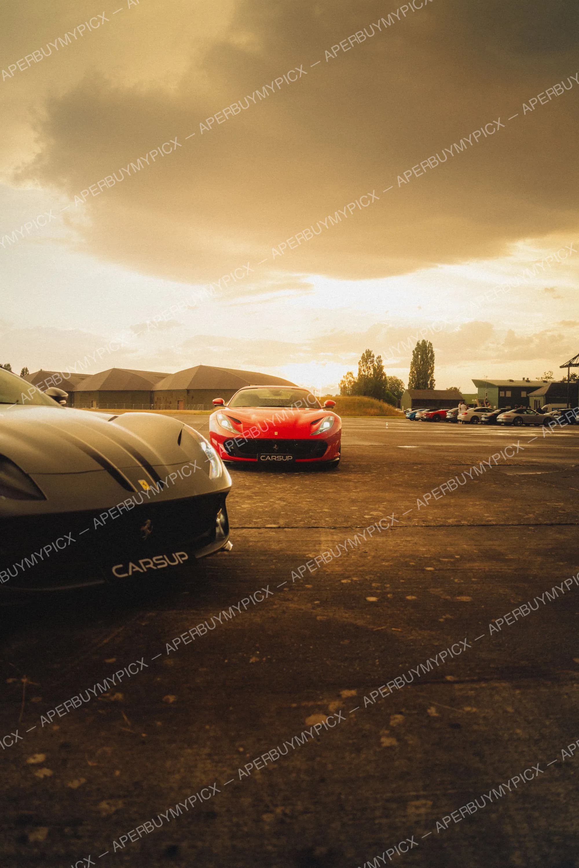 Deux Ferrari 812, une noire et une rouge, garées sur une piste d'aéroport au coucher de soleil avec des hangars et d'autres voitures en arrière-plan