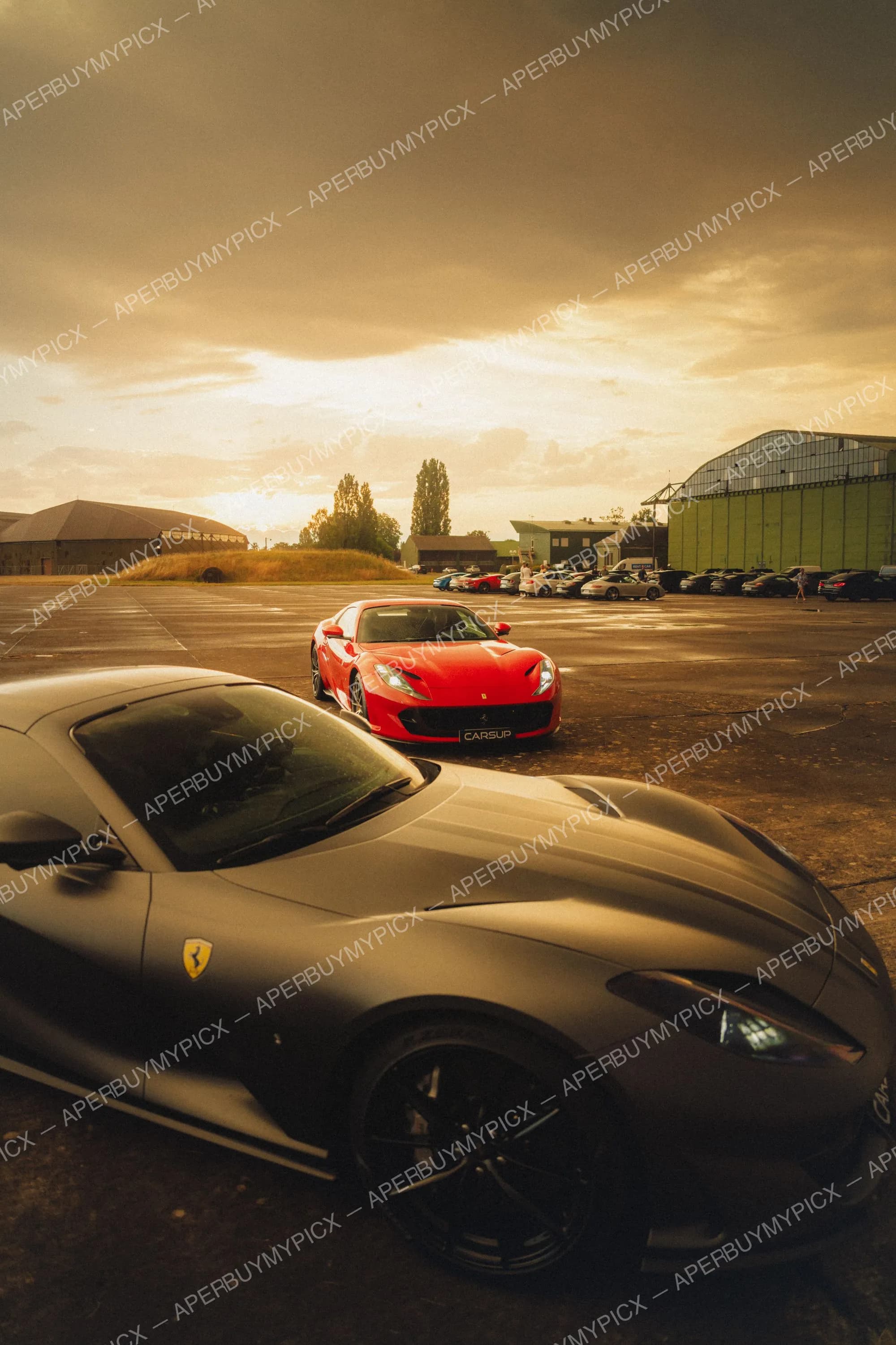 Une Ferrari 812 rouge et une Ferrari noire mat stationnées sur un ancien aérodrome au coucher du soleil, avec des hangars et d'autres voitures de sport en arrière-plan