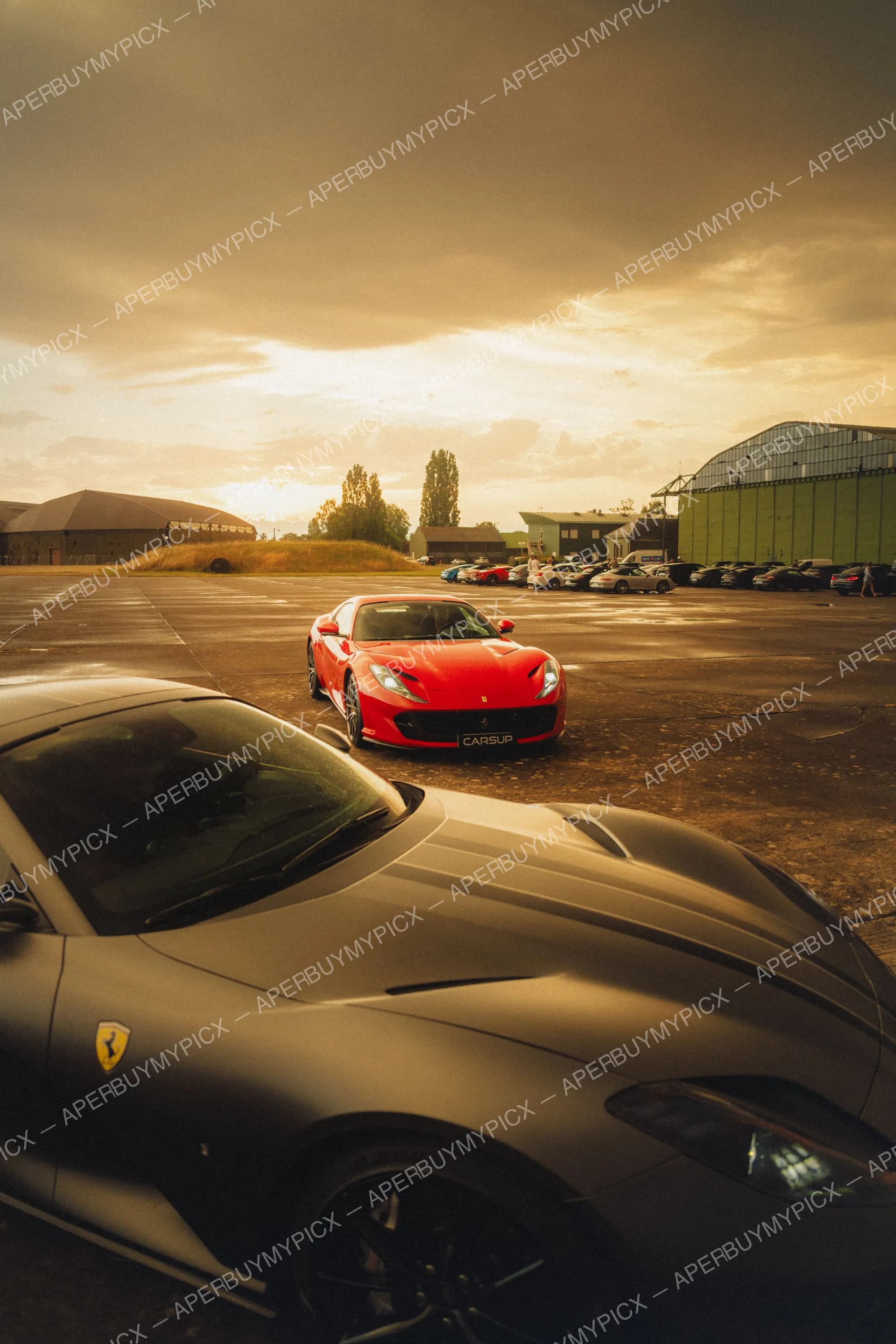 Ferrari 812 Superfast rouge garée sur un tarmac d'aérodrome au coucher de soleil, avec une Ferrari grise au premier plan, des hangars verts et une rangée de voitures de sport à l'arrière-plan sous un ciel doré nuageux.