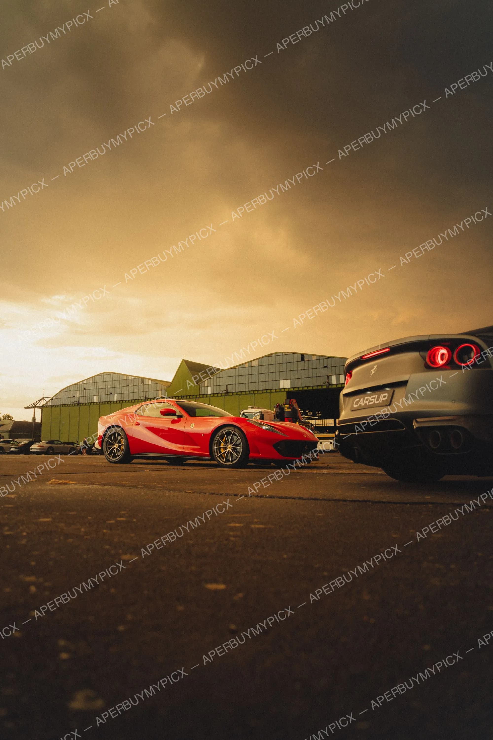 Ferrari 812 Superfast rouge garée à côté d'une Ferrari 812 grise vue de l'arrière, devant un hangar vert sous un ciel orageux et doré au coucher de soleil