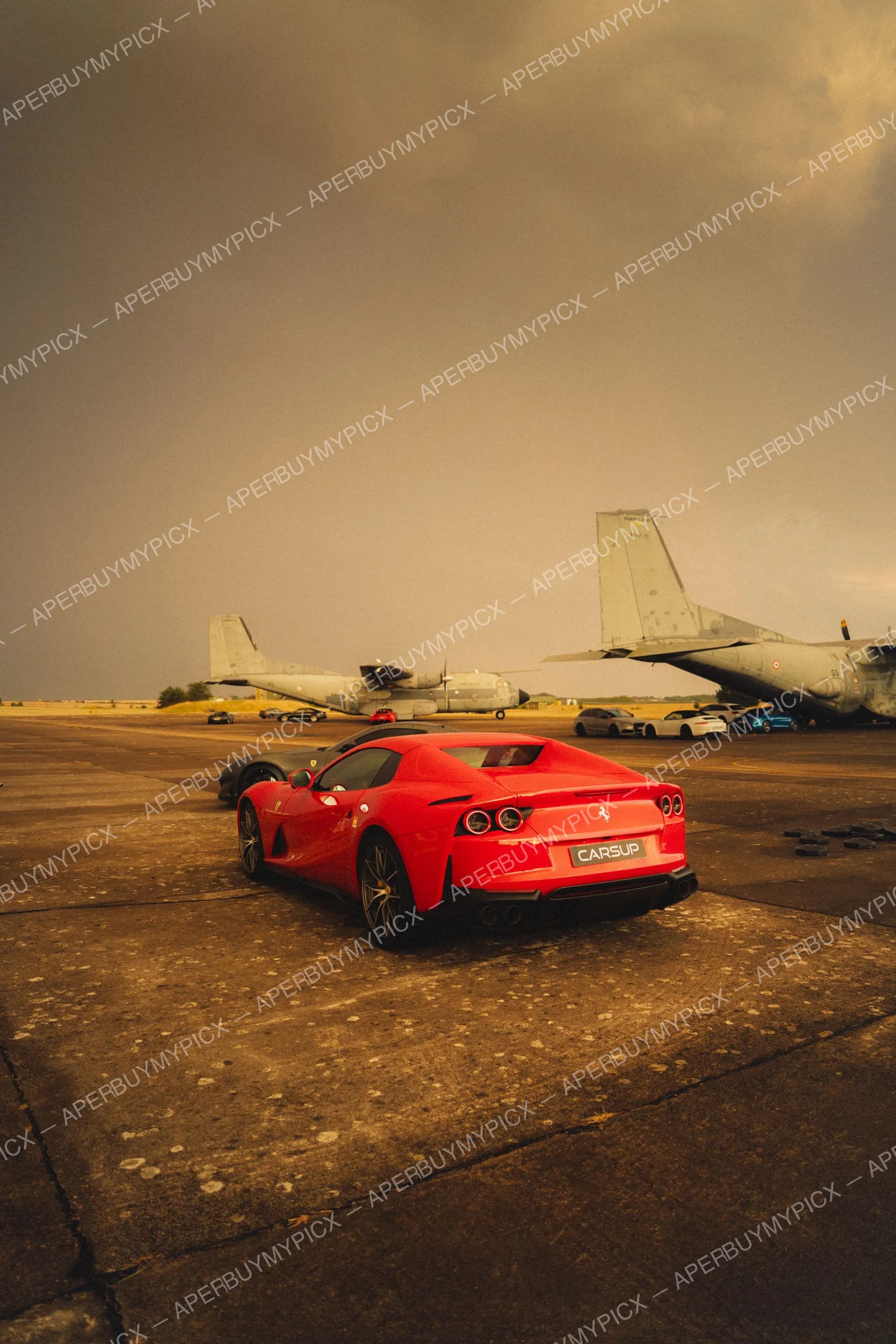 Ferrari 812 Superfast rouge vue de trois quarts arrière sur une piste d'aéroport militaire, avec des avions de transport gris et un ciel orageux en arrière-plan