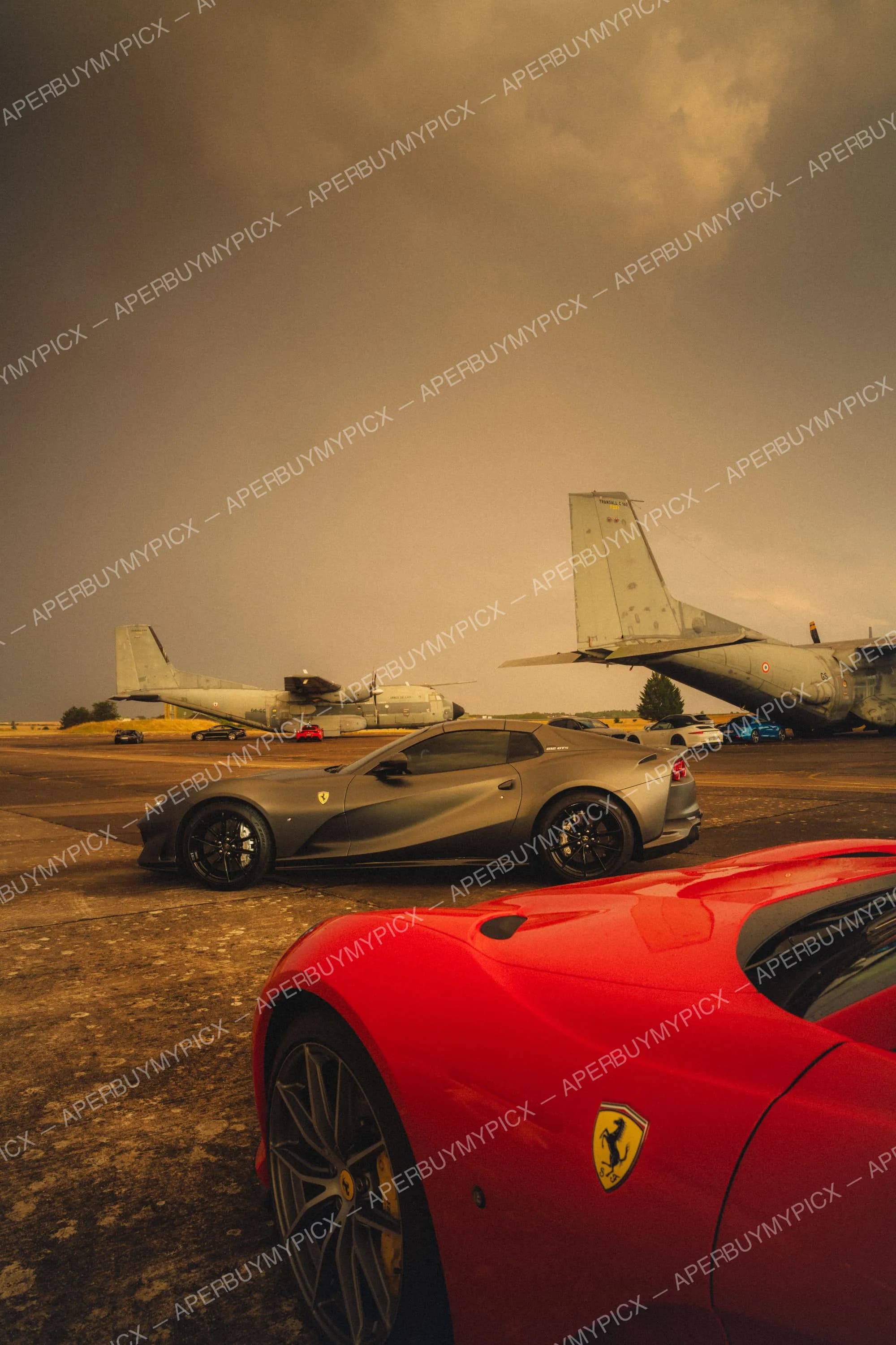 Ferrari 812 GTS grise mate et Ferrari rouge stationnées sur le tarmac d'une base aérienne, devant des avions de transport militaires sous un ciel orageux.